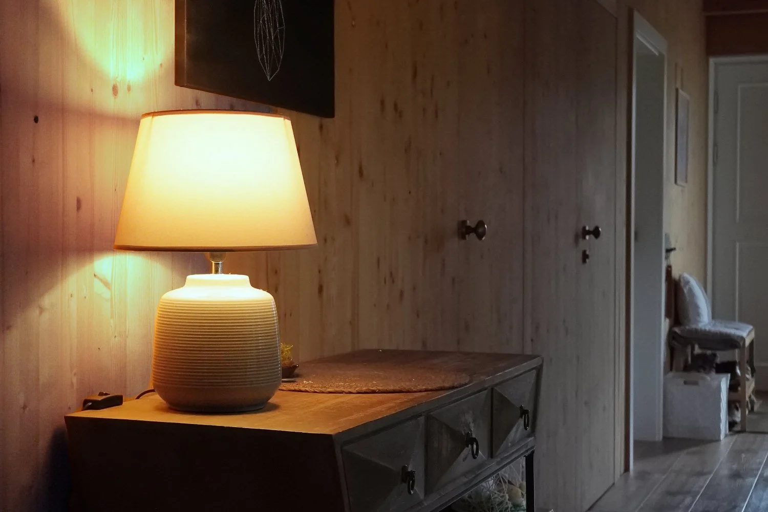 A moody interior scene of a lit table lamp in the foreground casting a warm glow down a dark wood-cladded hallway disappearing around a corner.