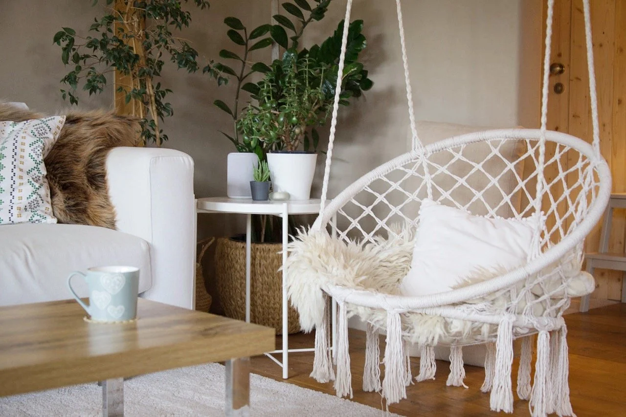 Cosy living room with a white sofa, decorated pillows, a wooden coffee table with a mug, a white hanging chair with a cushion and fluffy throw, indoor plants on a small white table, and wooden flooring with a wall and staircase in the background.