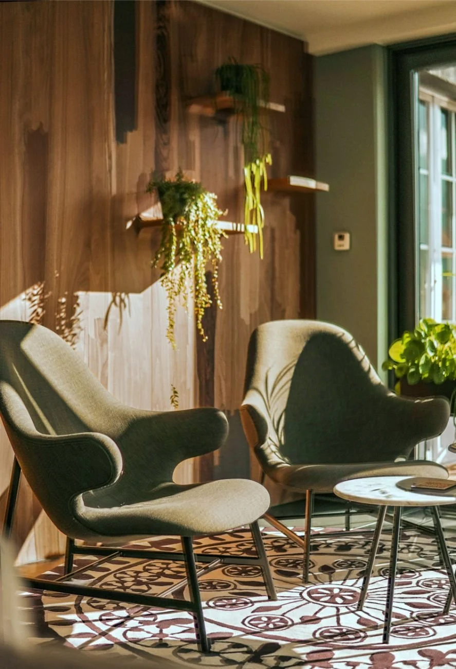 • A portrait shot of a safe seating area featuring a dark wood clad wall, two mossy green high-back upholstered armchairs, plants, and soft ambient sunlight.
