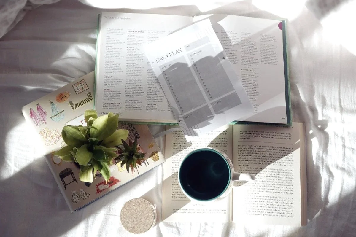 Cozy bedroom scene with open books spread on the bed, a small potted plant, and a mug, creating a relaxed and inviting reading nook.