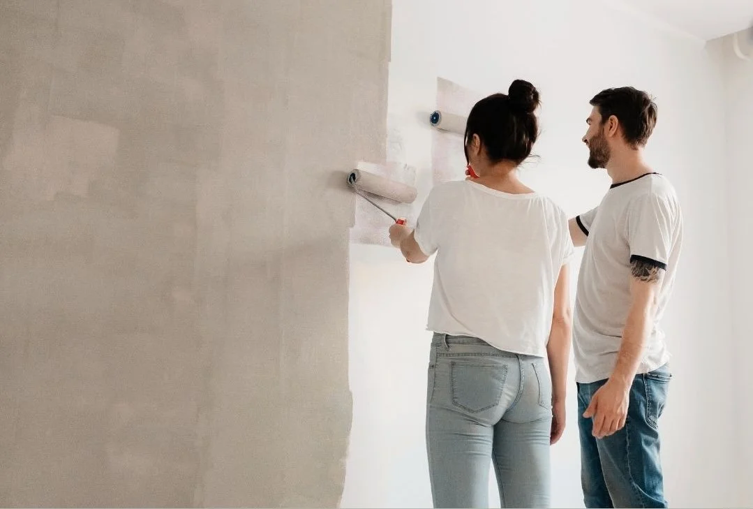 Man and woman painting their interior wall with beige paint — choosing low-VOC paints can help improve air quality.