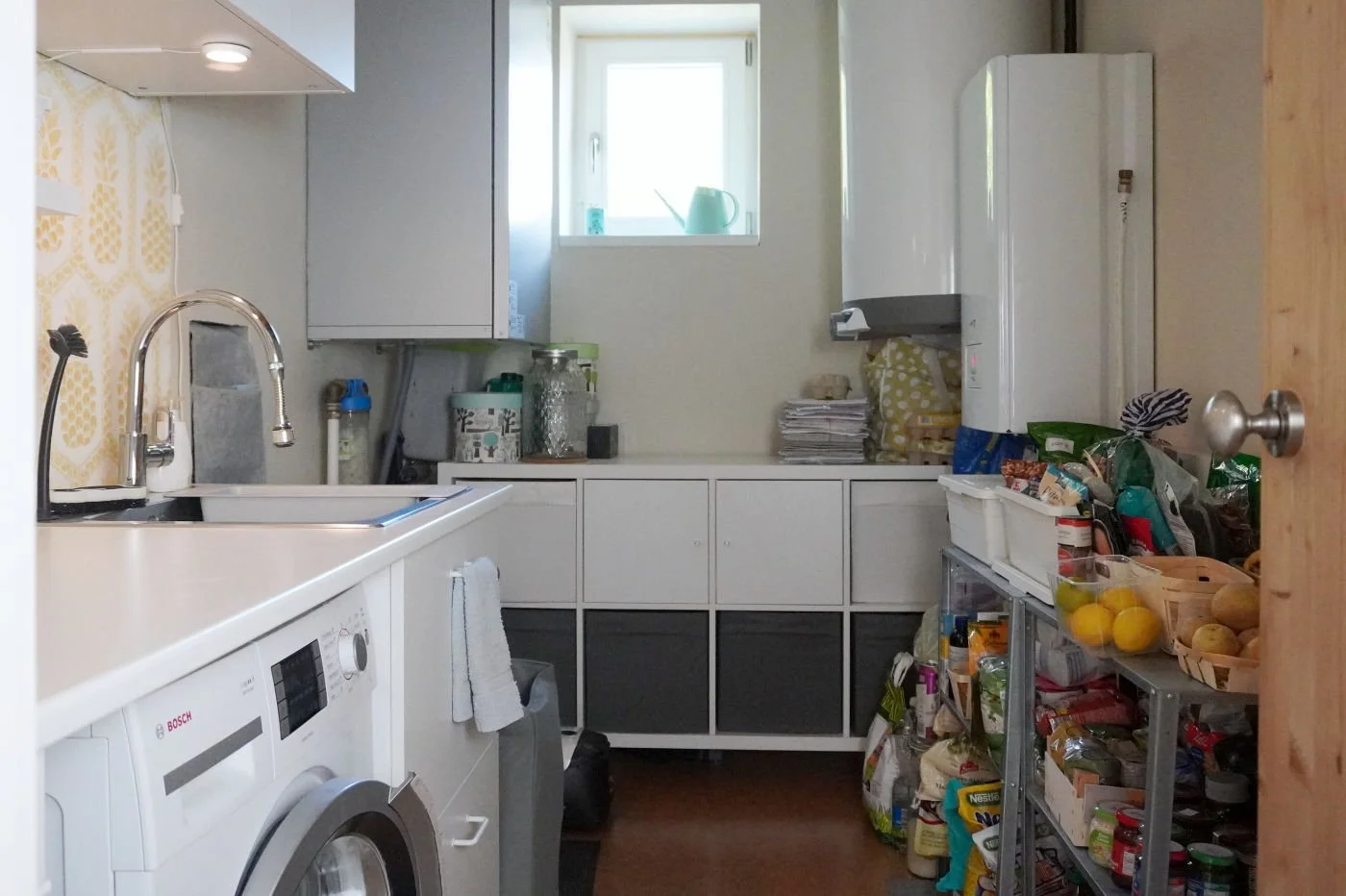 Wide shot of a functional small utility room layout with a laundry zone on the left, open pantry shelves on the right, and home technical equipment like a boiler and MVHR unit.