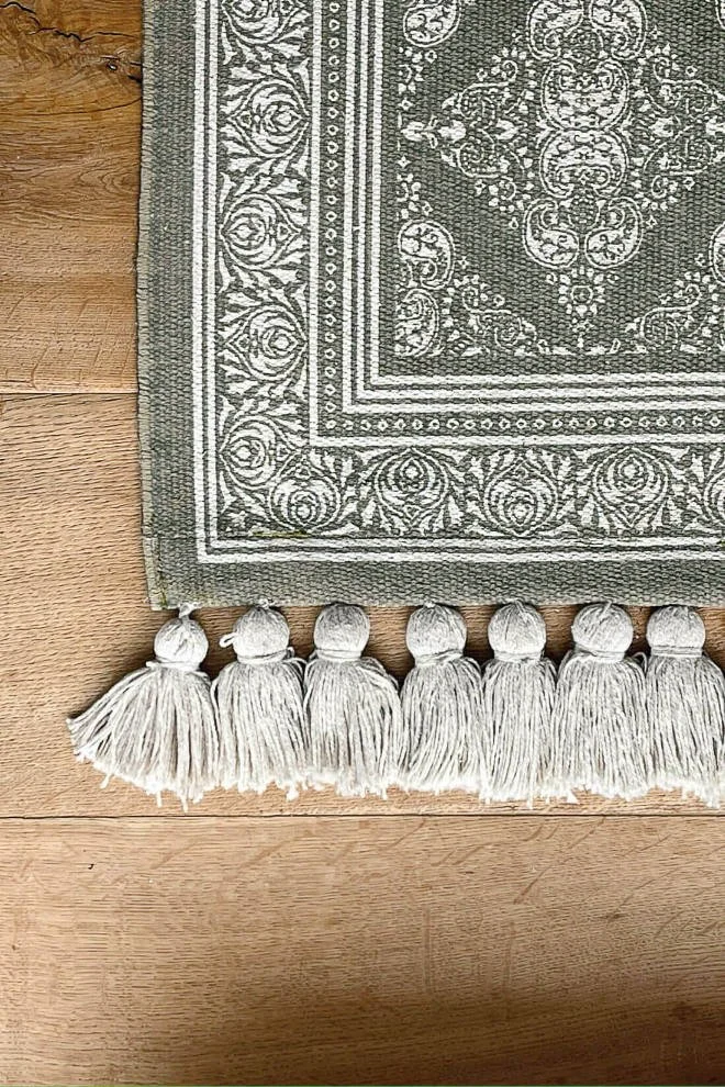 Close-up of a geometric kitchen runner in muted green and cream tones on a wooden floor with thick tasselled edges.