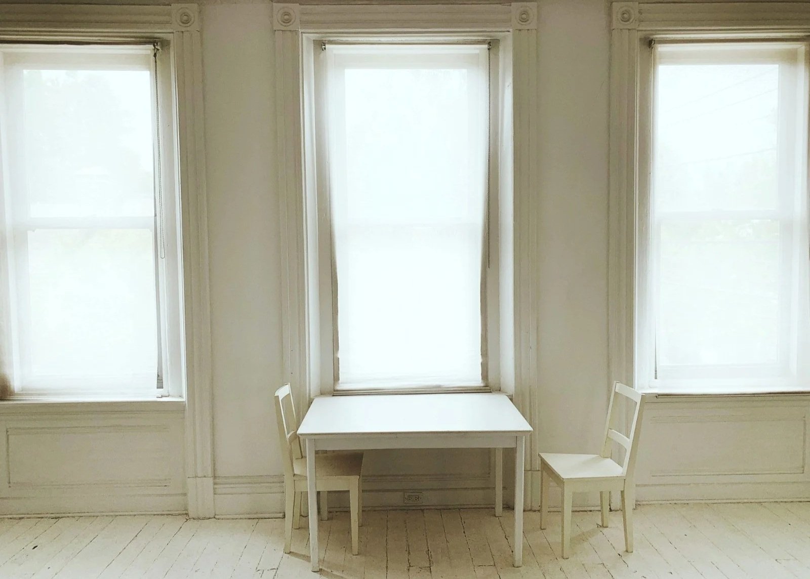 Bright white studio apartment with large windows and sunlight, showing a simple dining table and two chairs.