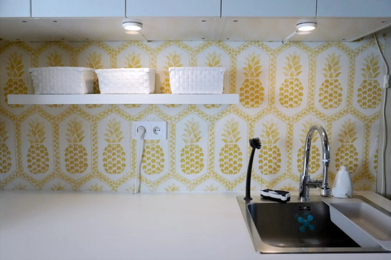 Front-on view of a utility room countertop with a large stainless steel sink, under-cabinet spotlights, a white wall shelf holding three small woven baskets, and gold pineapple wallpaper.