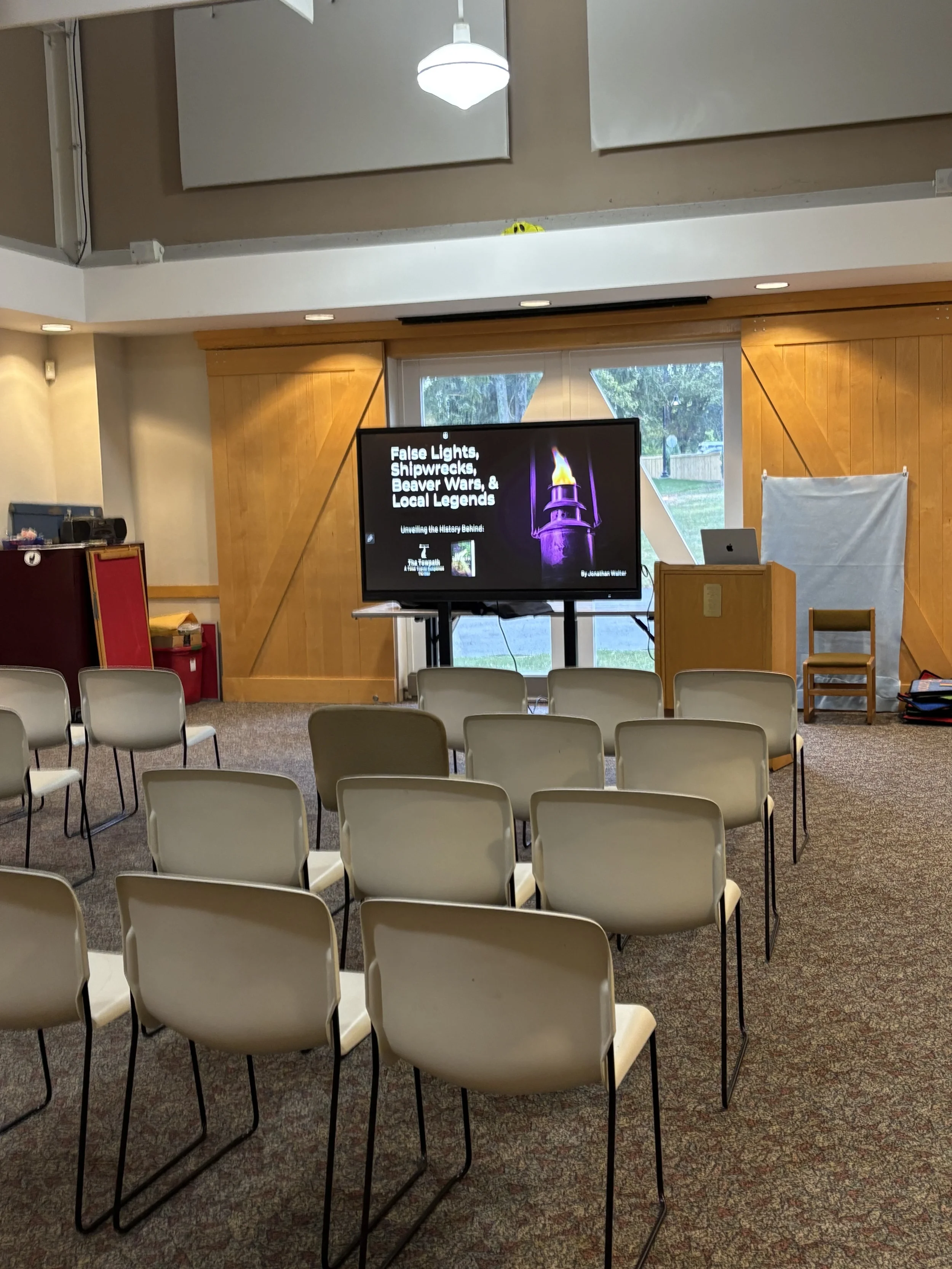 An empty classroom or lecture hall with multiple rows of beige chairs facing a large screen displaying a presentation titled 'False Lights, Shipwrecks, Beaver Wars, & Local Legends.' The room has a high ceiling with white acoustic panels, wooden wall
