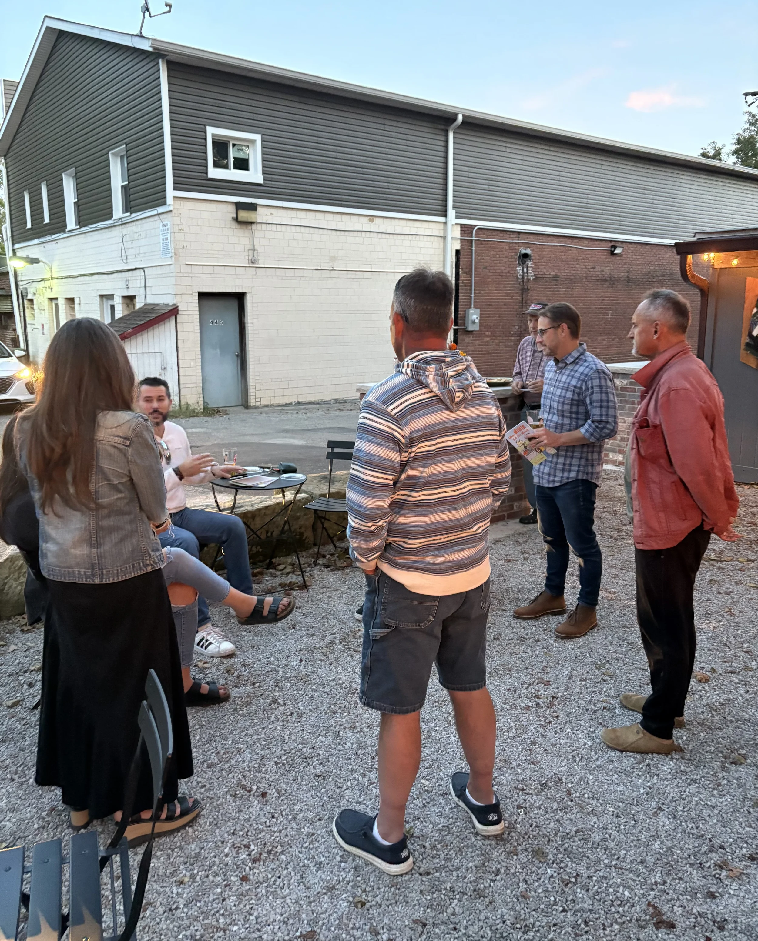 Group of people socializing outdoors at dusk, standing and sitting near a small table, in an urban setting.