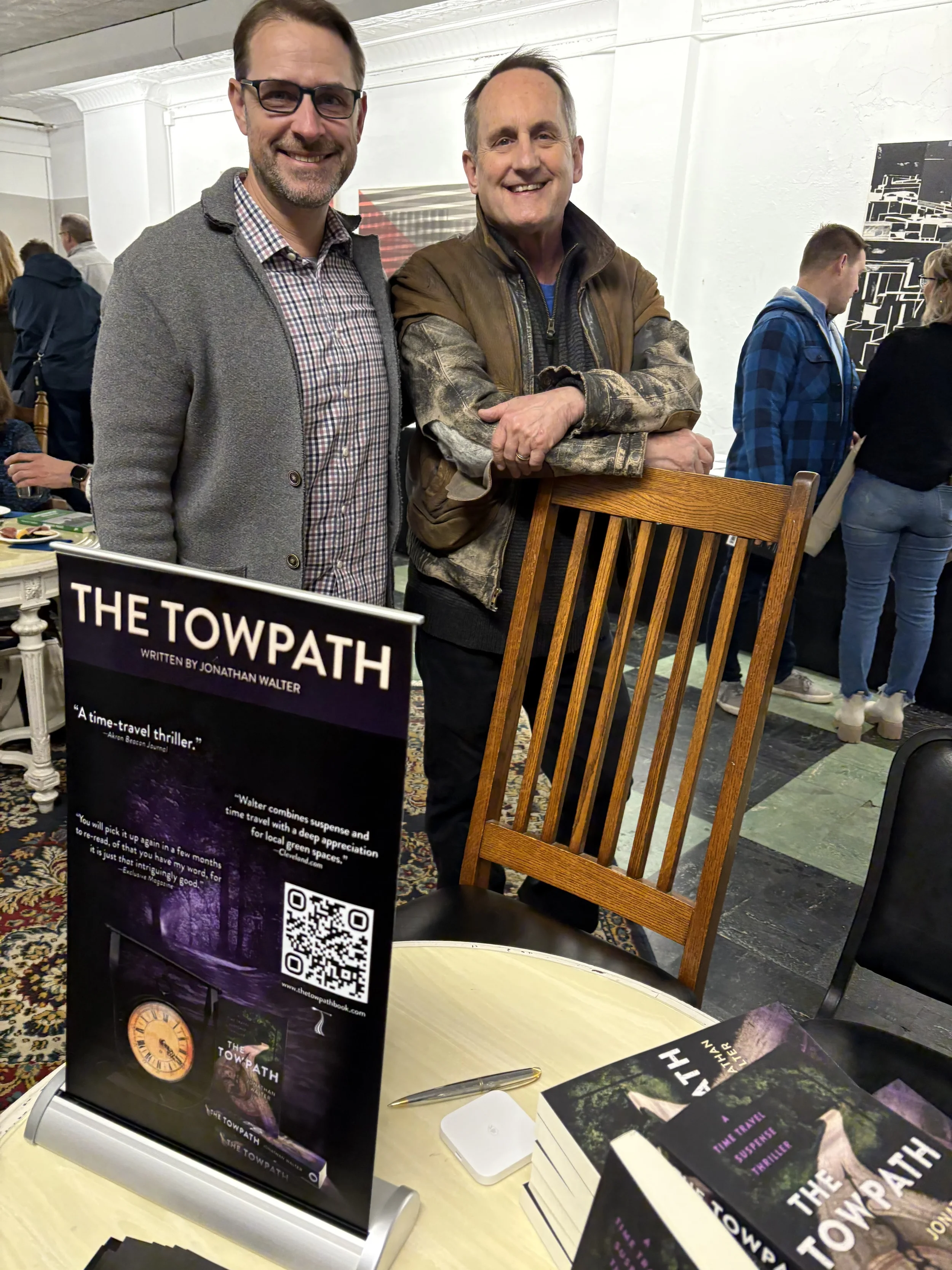 Two men standing behind a table promoting a book titled 'The Towpath.' One man is wearing glasses and a gray blazer, and the other is wearing a brown leather jacket with camouflage sleeves. There are people in the background at an indoor event.
