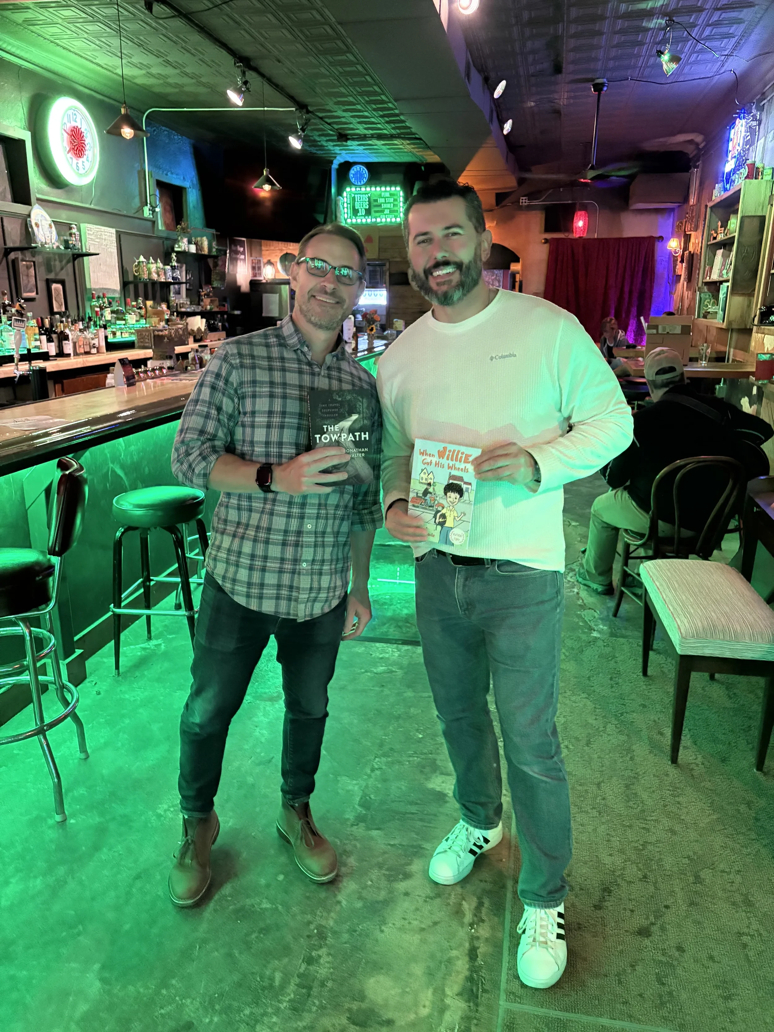 Two men standing inside a bar or pub, holding books and smiling at the camera. One man is wearing a plaid shirt and glasses, the other a white long sleeve shirt and sneakers. The bar has neon signs, shelves with bottles, and a few patrons sitting at
