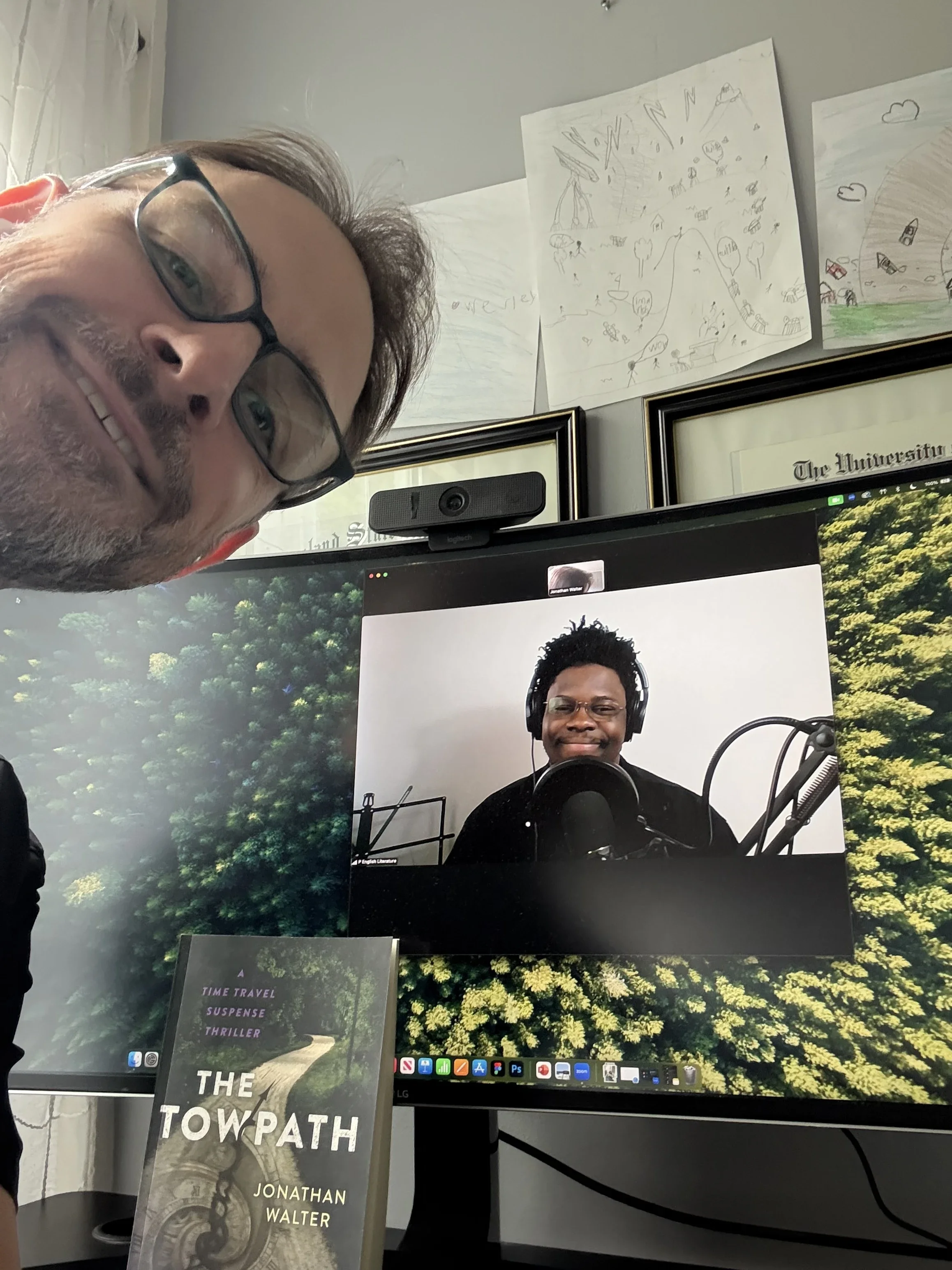 A man with glasses and a beard taking a selfie in front of a computer monitor displaying a video call with another man who is wearing headphones and speaking into a microphone. There are children's drawings and framed certificates on the wall behind