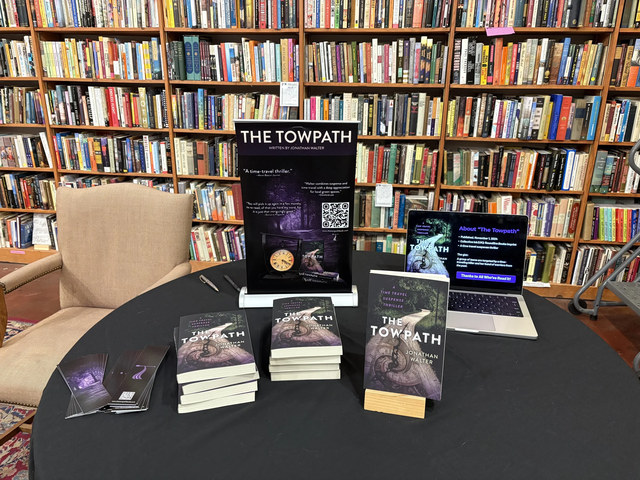 A table set up for a book signing event with copies of 'The Towpath' by Jonathan Walter, a promotional poster, and a laptop displaying information about the book, surrounded by bookshelves filled with various books.