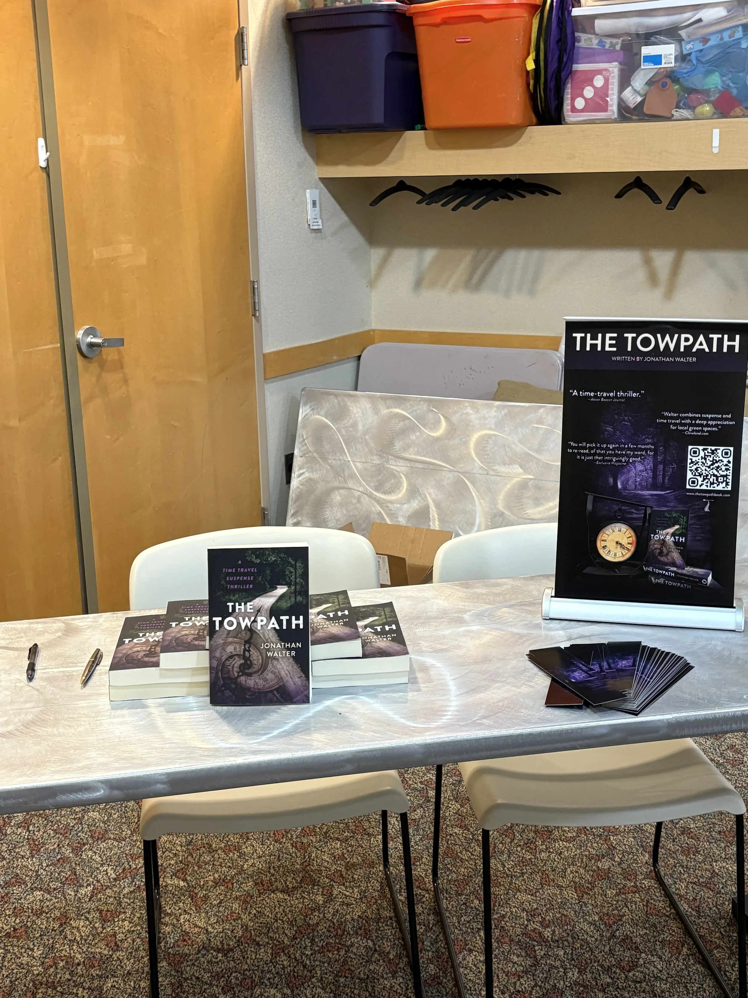 Table set up with copies of the book "The Towpath" by Jonathan Walter, promotional materials, and pens, for a book signing or display event, in a room with beige walls, wooden door, and colorful storage bins on shelves.