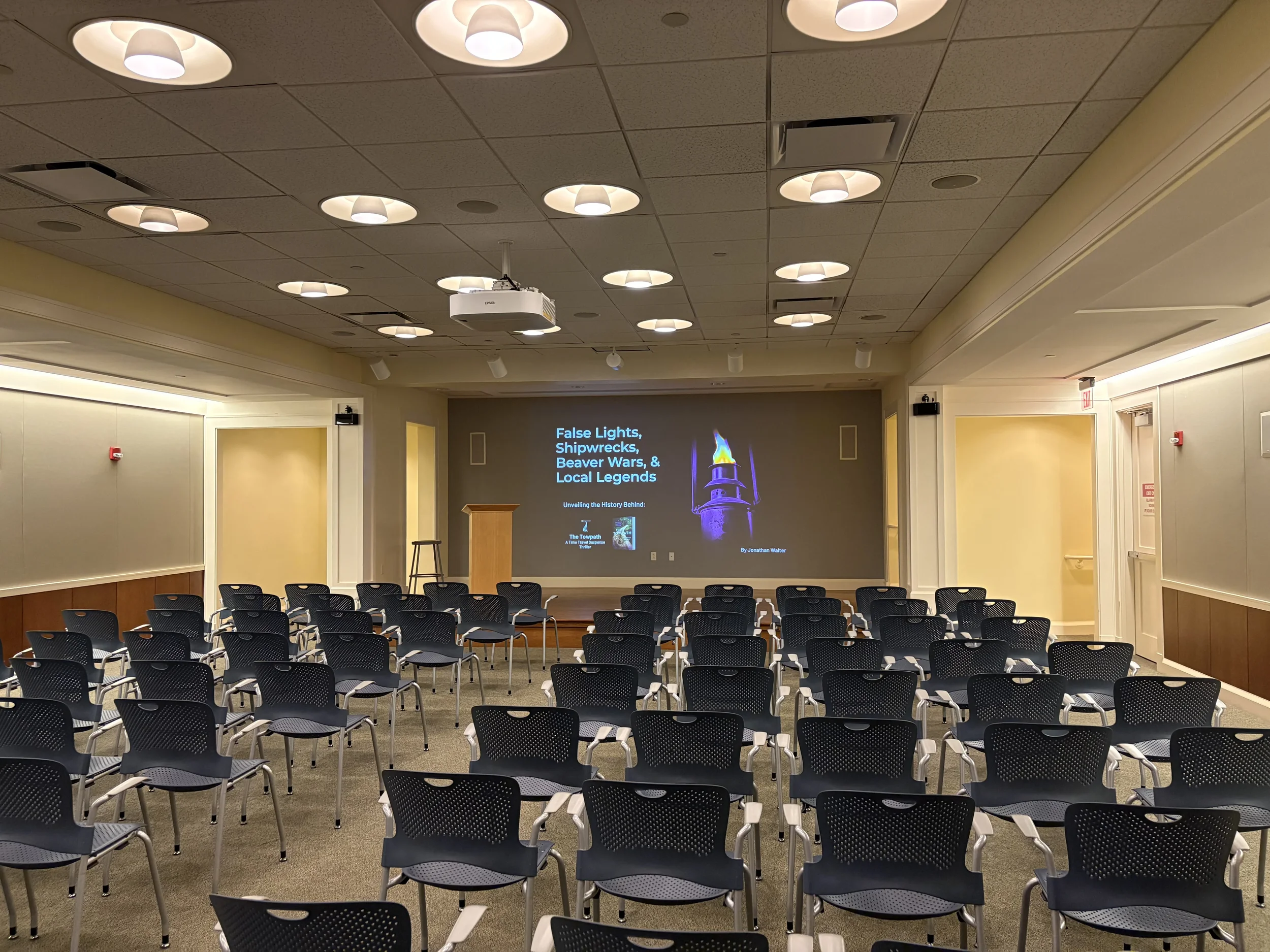 An empty conference room with rows of black chairs facing a projection screen displaying a presentation titled 'False Lights, Shipwrecks, Beaver Wars, & Local Legends' by Jonathan Walter, with a graphic of a lighthouse.