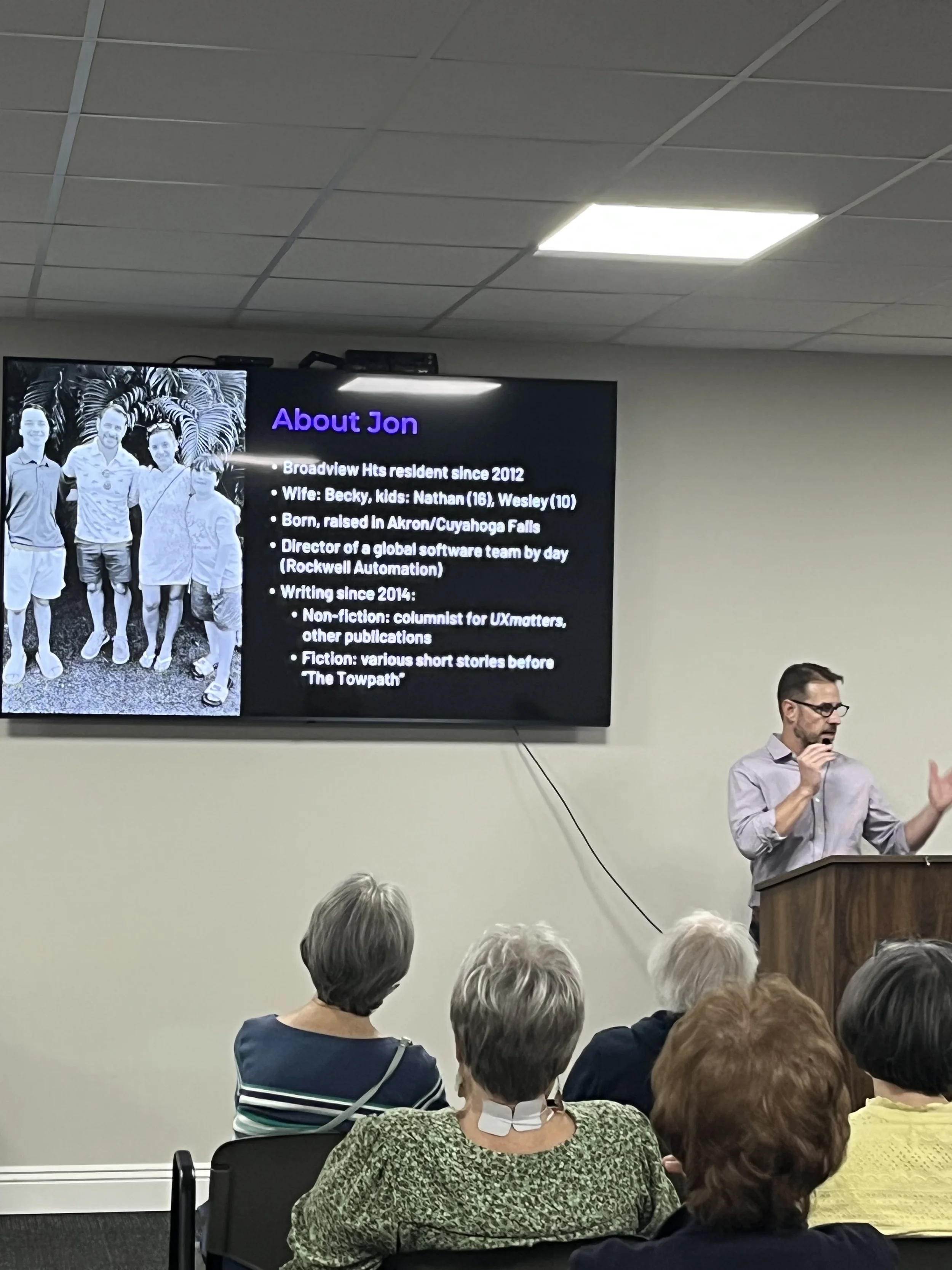 A man giving a presentation in front of an audience of mostly older women. A large screen behind him displays a black and white photo of four young men and some text introducing Jon, including personal background and writing achievements.