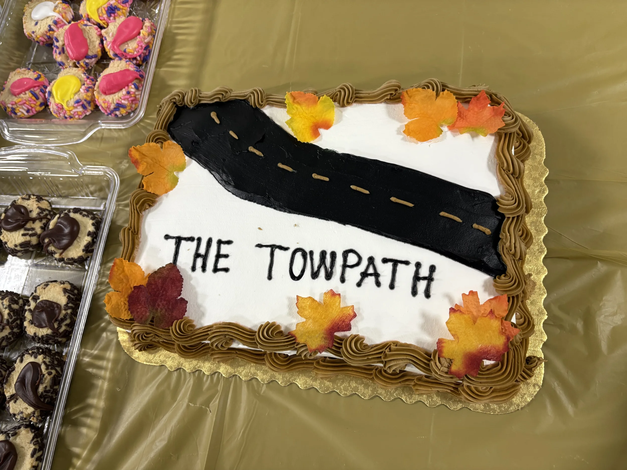 A decorated birthday cake with the words "The Towpath" and a road design, surrounded by fall leaves, on a table with other desserts.