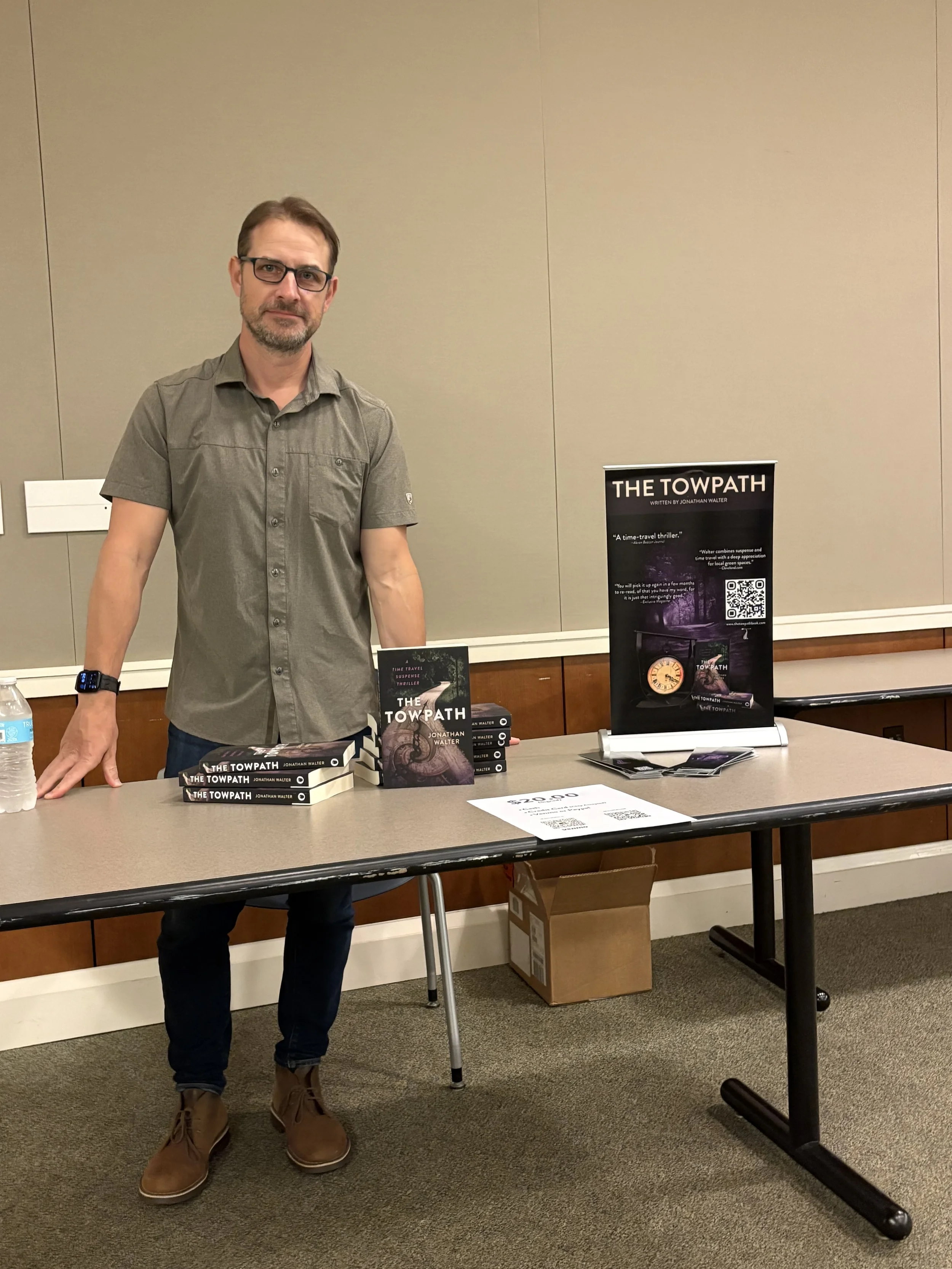 Author Jonathan Walter standing behind a table with multiple copies of his book, 'The Towpath,' at a book signing or promotional event in a conference room.