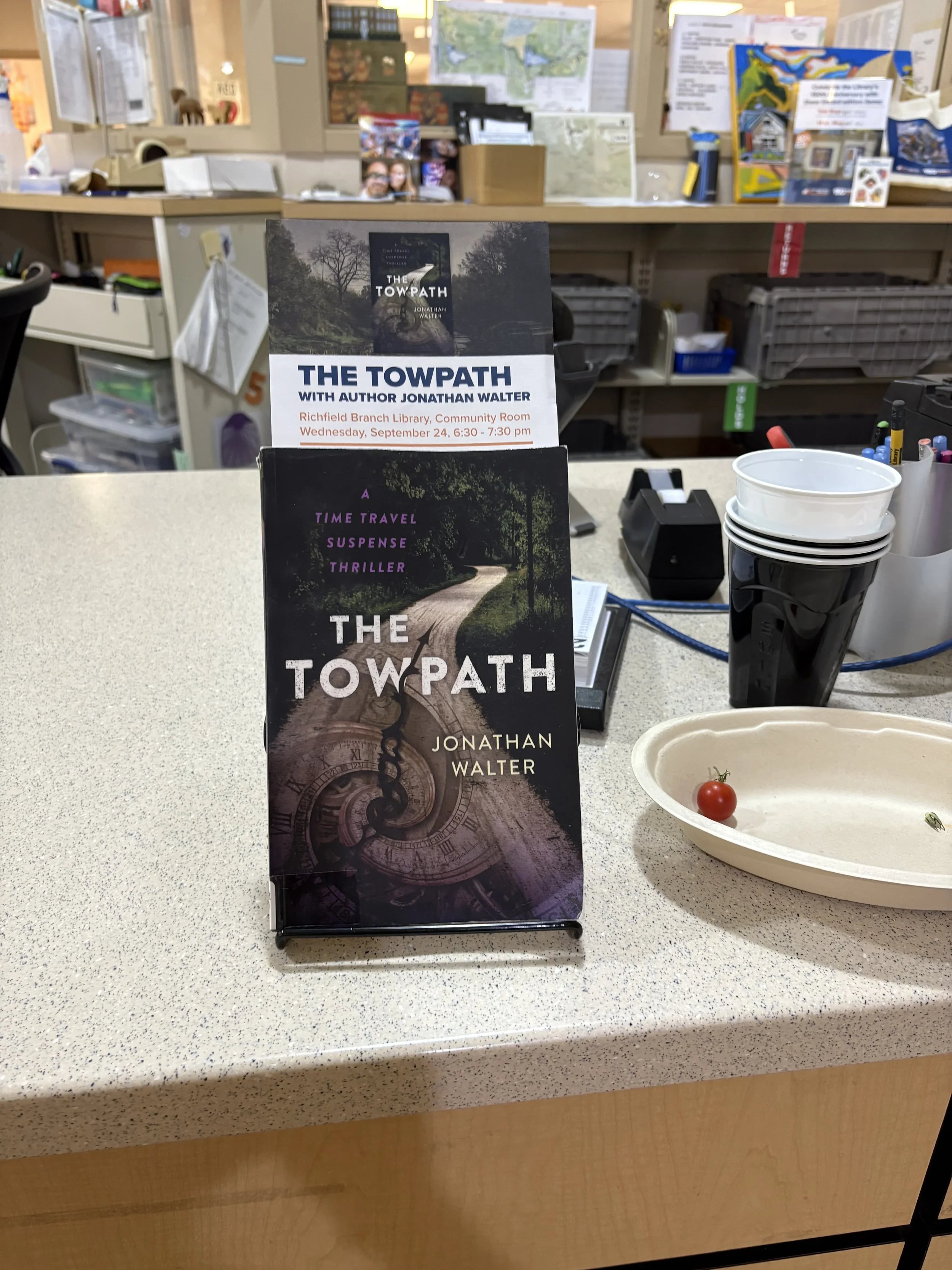 A brochure for the book "The Towpath" by Jonathan Walter displayed on a counter at a library or bookstore, with promotional material for an author event on September 24. Behind the brochure, there are stacks of cups, pens, and a cash register, sugges