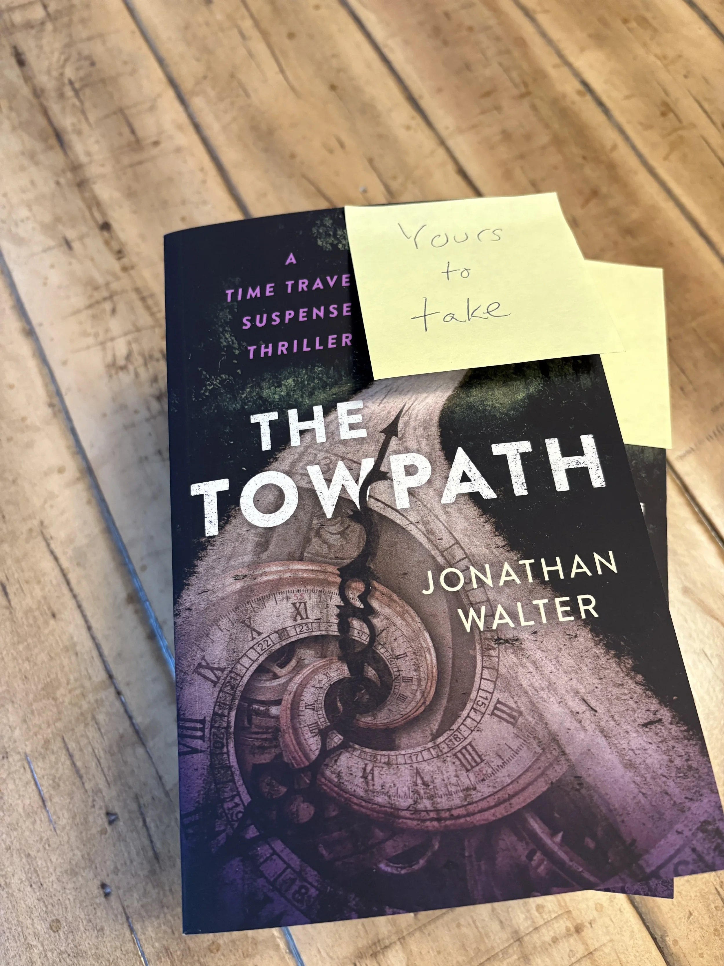 A paperback novel titled 'The Towpath' by Jonathan Walter, with a yellow sticky note on top that says 'Yours to take'. The book cover features a spiral of clock faces on a dark background.