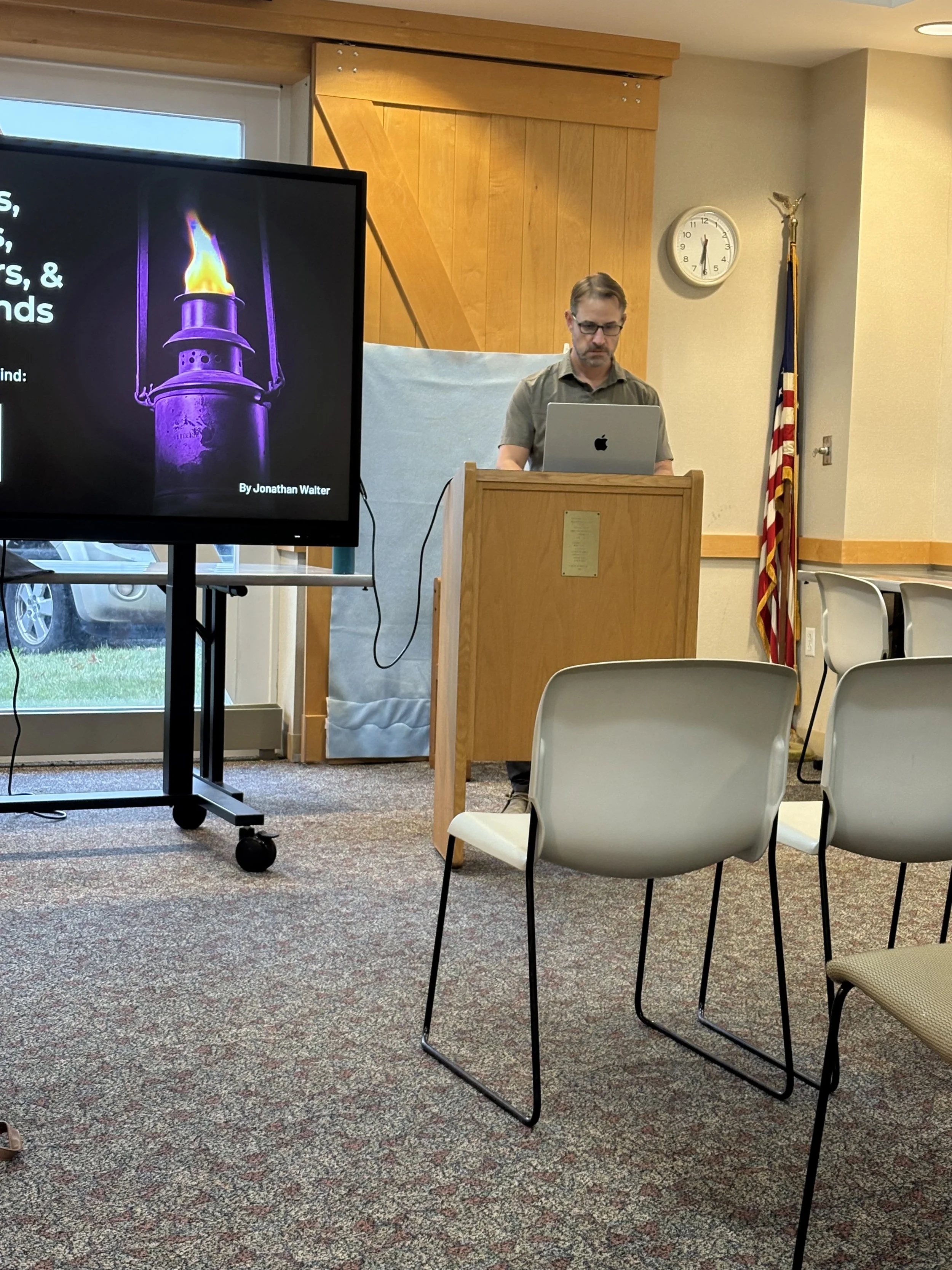 A man standing at a wooden podium in a room, looking at a silver laptop, with a large screen to his left displaying a presentation slide with a purple lantern and text. There are empty chairs arranged in rows in front of him, and a clock, American fl