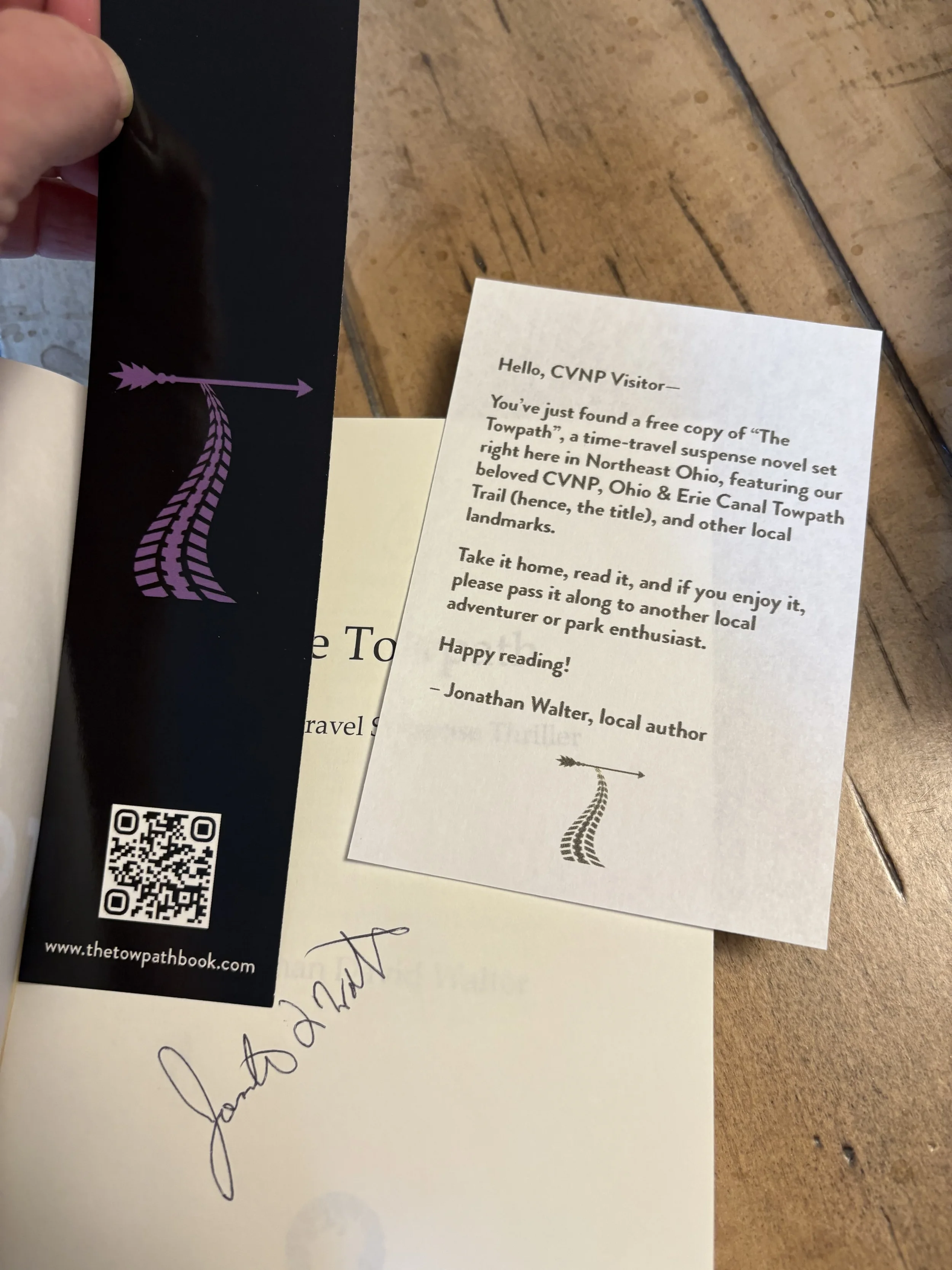 Close-up of a hand holding a bookmark with purple and black arrow and trail graphic, next to a white paper with black text and a printed signature on a wooden surface.