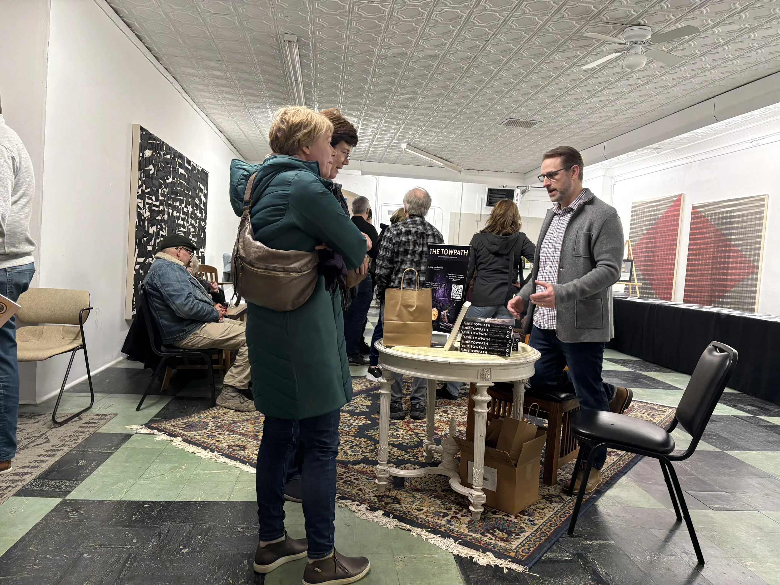 People at an indoor event, with a man explaining something to two women at a display table with books and a sign titled 'The Towpath'; others are seated or standing around in the background, with various artwork on the walls.