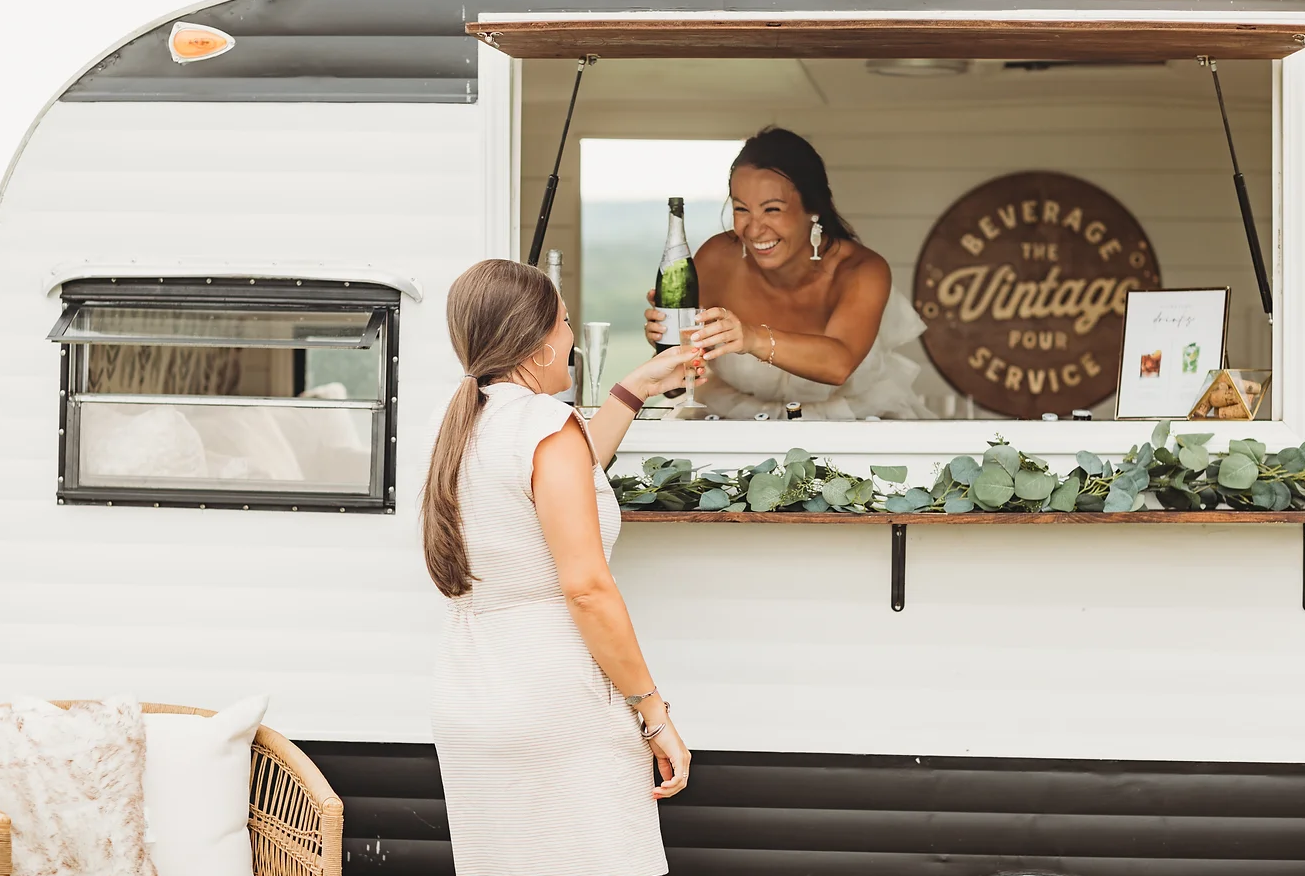 A woman in a white dress is at a food truck, receiving a bottle of champagne and a glass from a smiling woman inside the truck. The truck has a eucalyptus decoration and a sign that reads 'Beverage The Vintage Pour Service.'