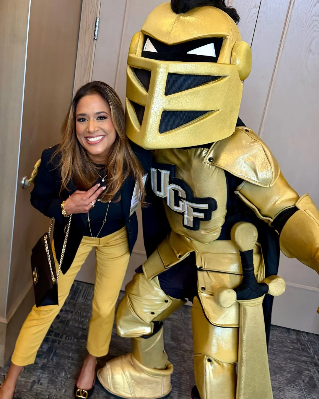 Woman with long brown hair wearing a black blazer, light khaki pants, and a conference badge standing next to a person in a large, gold University of Central Florida (UCF) knight costume giving a thumbs-up inside a building with large windows and a vending machine in the background.