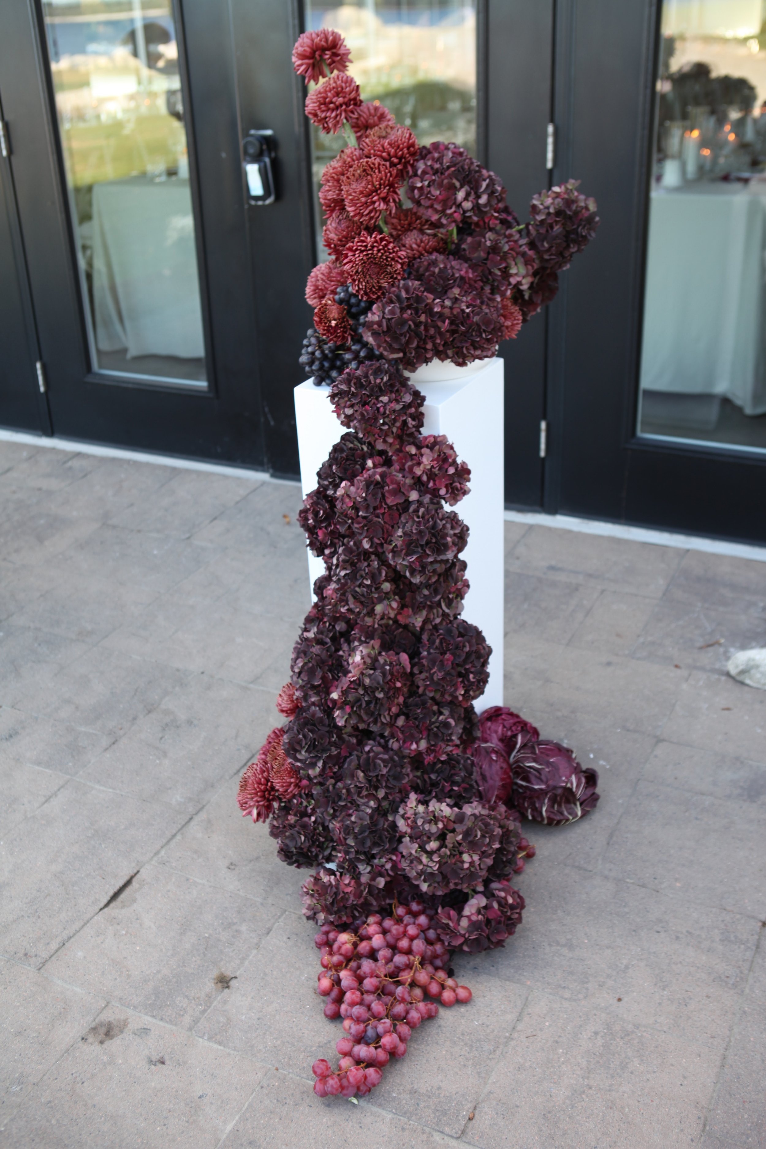 A floral arrangement of dark red and purple flowers including hydrangeas, dahlias, and grapes on a white pedestal outside a building with glass doors.