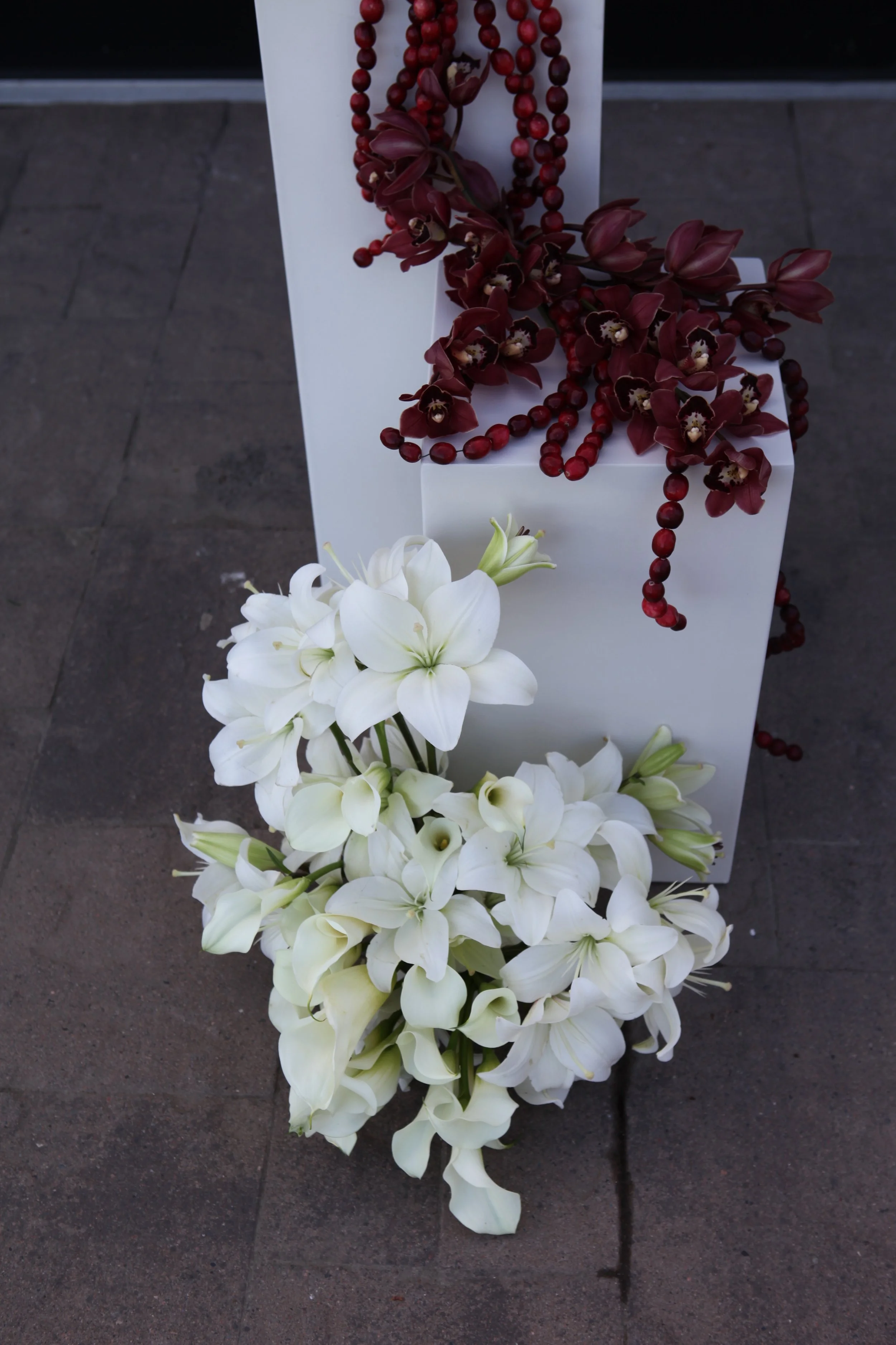 White lilies and burgundy orchids with beads on a white stand on a dark gray floor.