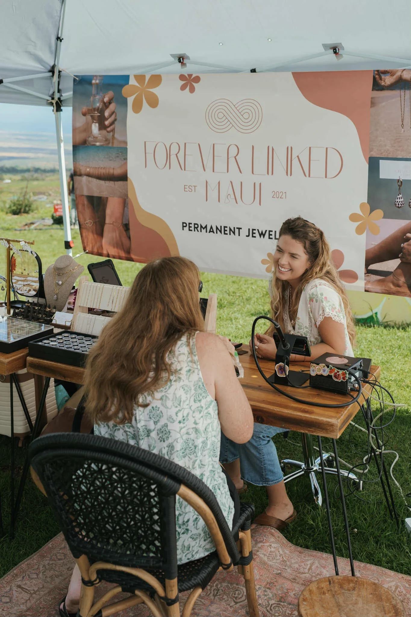 Image of the owner chatting with a client at her booth with products displayed on the table and a Forever Linked Maui banner behind them.