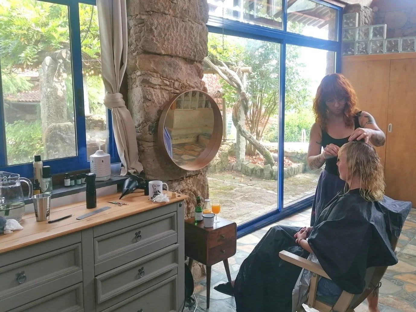 A woman sitting in a salon chair getting her hair done by a hairstylist in a bright room with large glass windows, stone walls, and wooden furniture.
