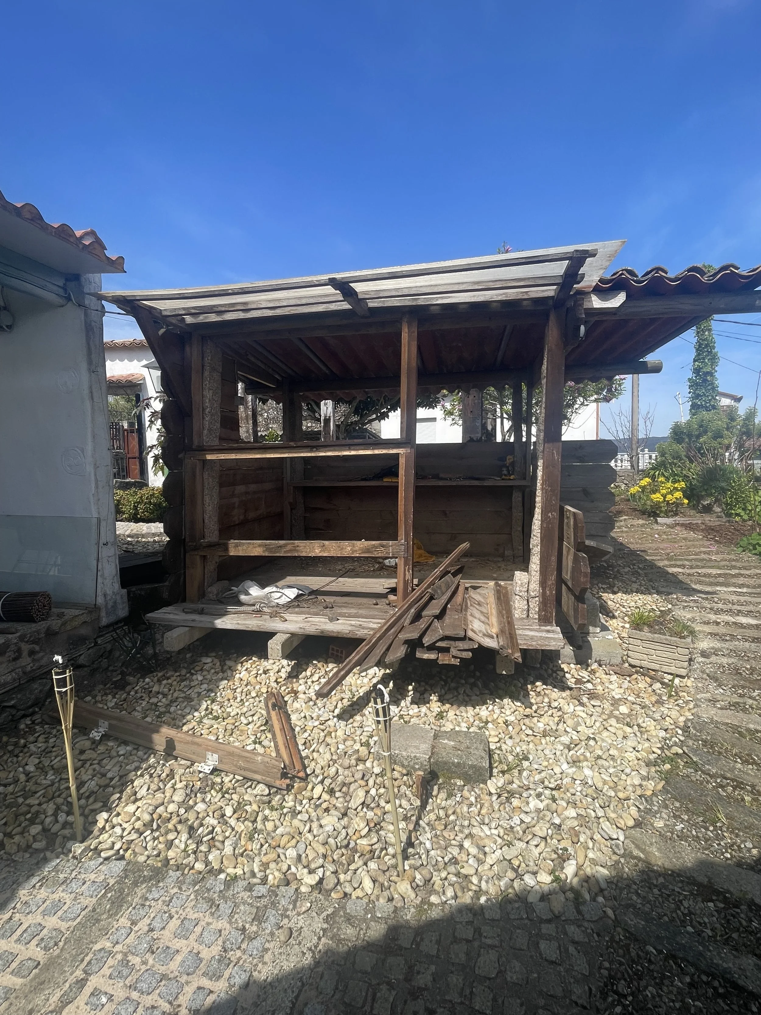 A damaged wooden structure with a sloped roof and open sides, surrounded by a gravel ground and nearby plants, under a clear blue sky.