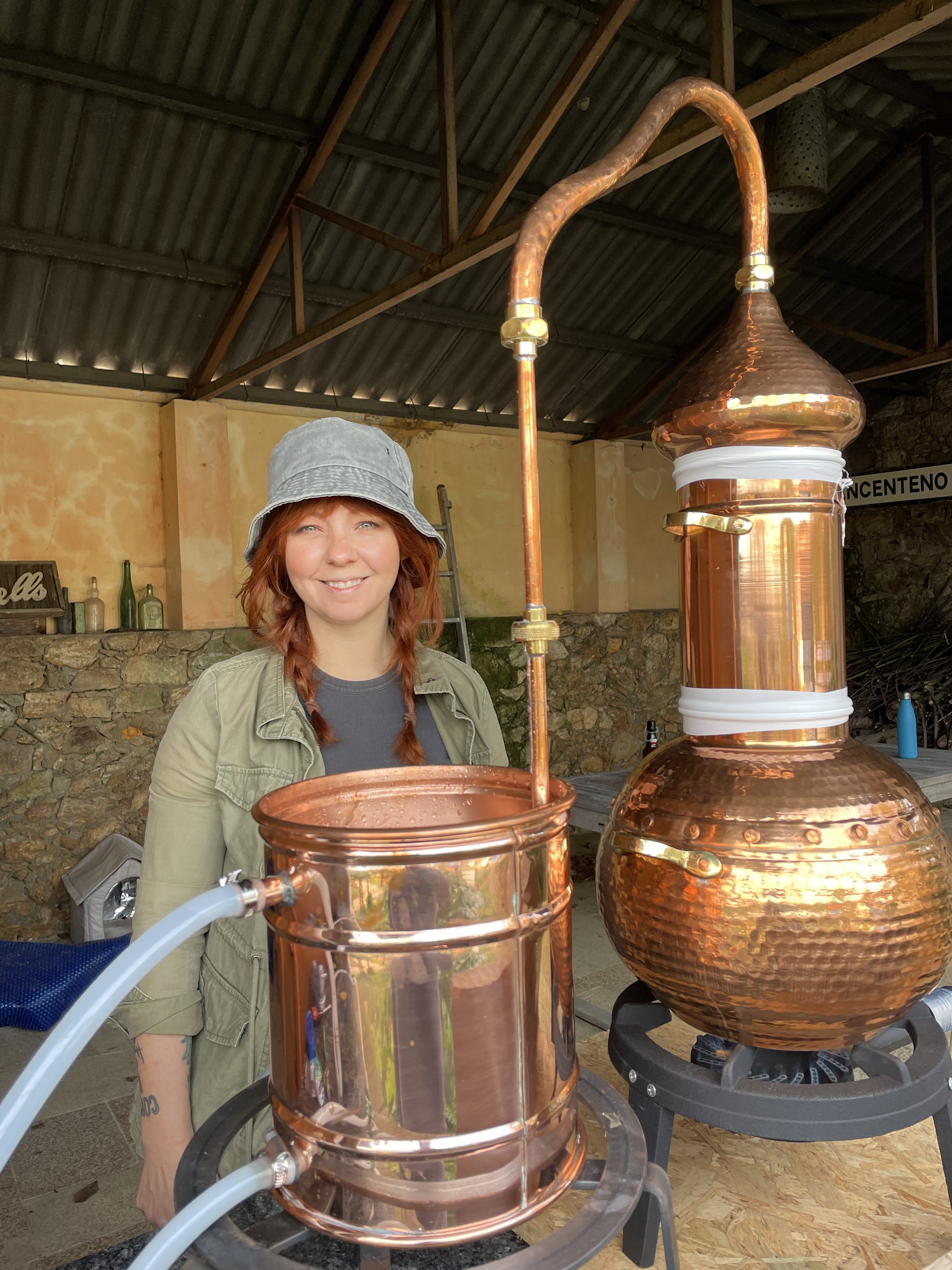 A woman with red hair, wearing a gray hat and green jacket, stands beside a large copper still in an indoor setting with stone walls and a metal roof, smiling at the camera.