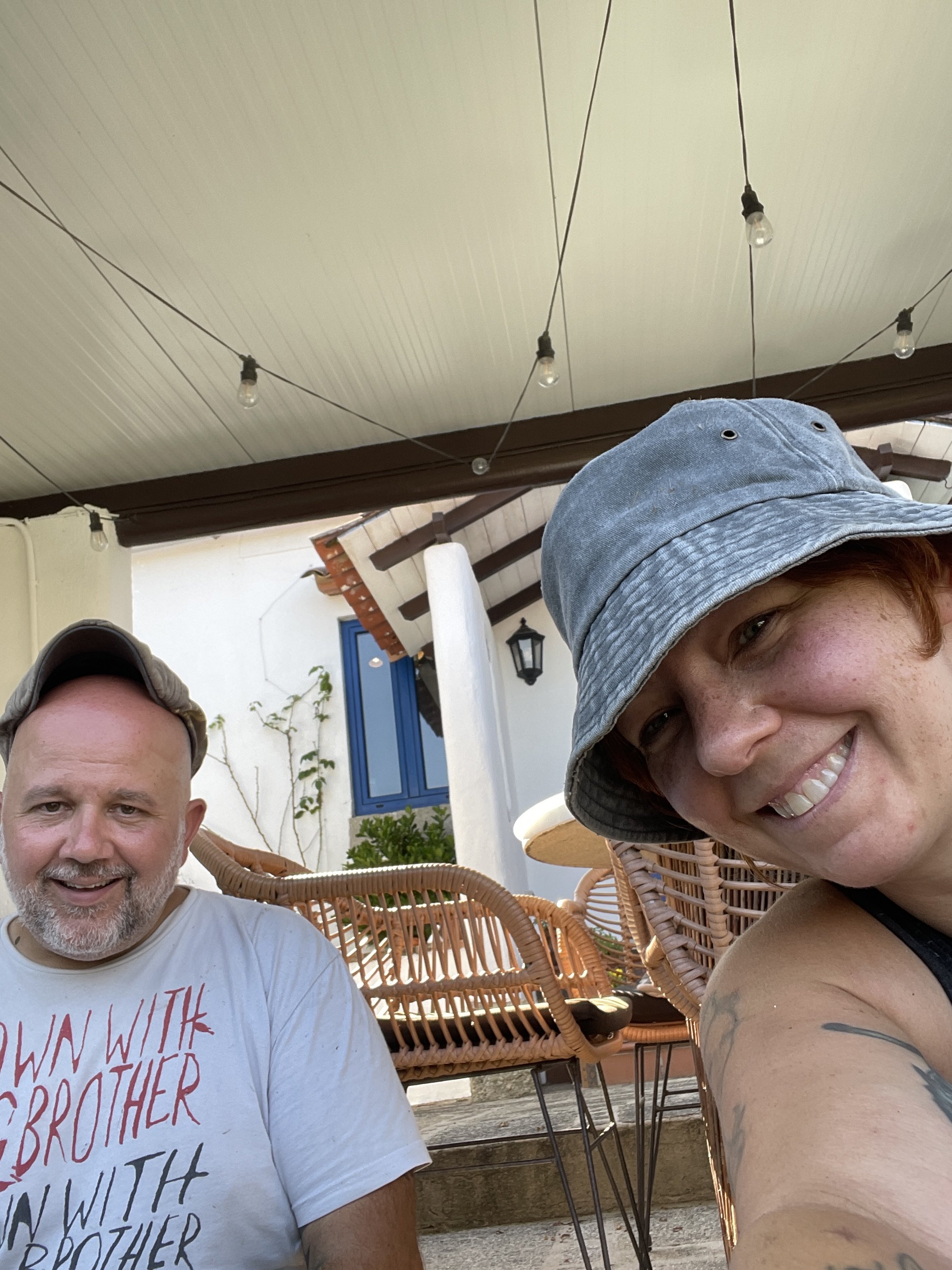 A man and a woman taking a selfie outdoors. The man has a beard, wearing a light t-shirt and a cap, smiling. The woman has short hair, wearing a bucket hat, smiling. There are wicker chairs and a building with a blue window in the background, with string lights hanging overhead.