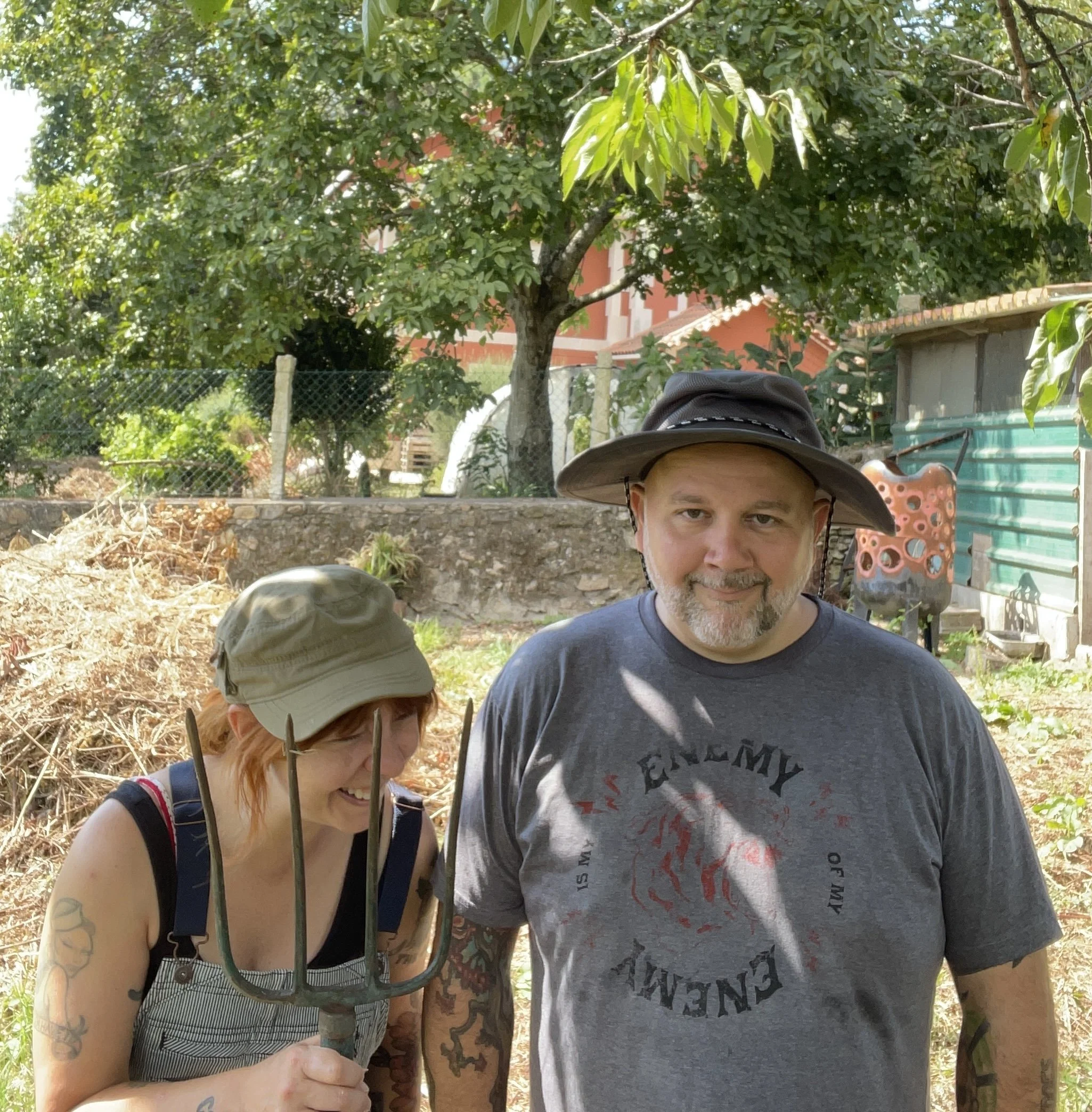 A woman and man outdoors in a garden, both wearing hats. The woman is holding a gardening fork and laughing, while the man has a slight smile. They are surrounded by trees, plants, and garden decorations.