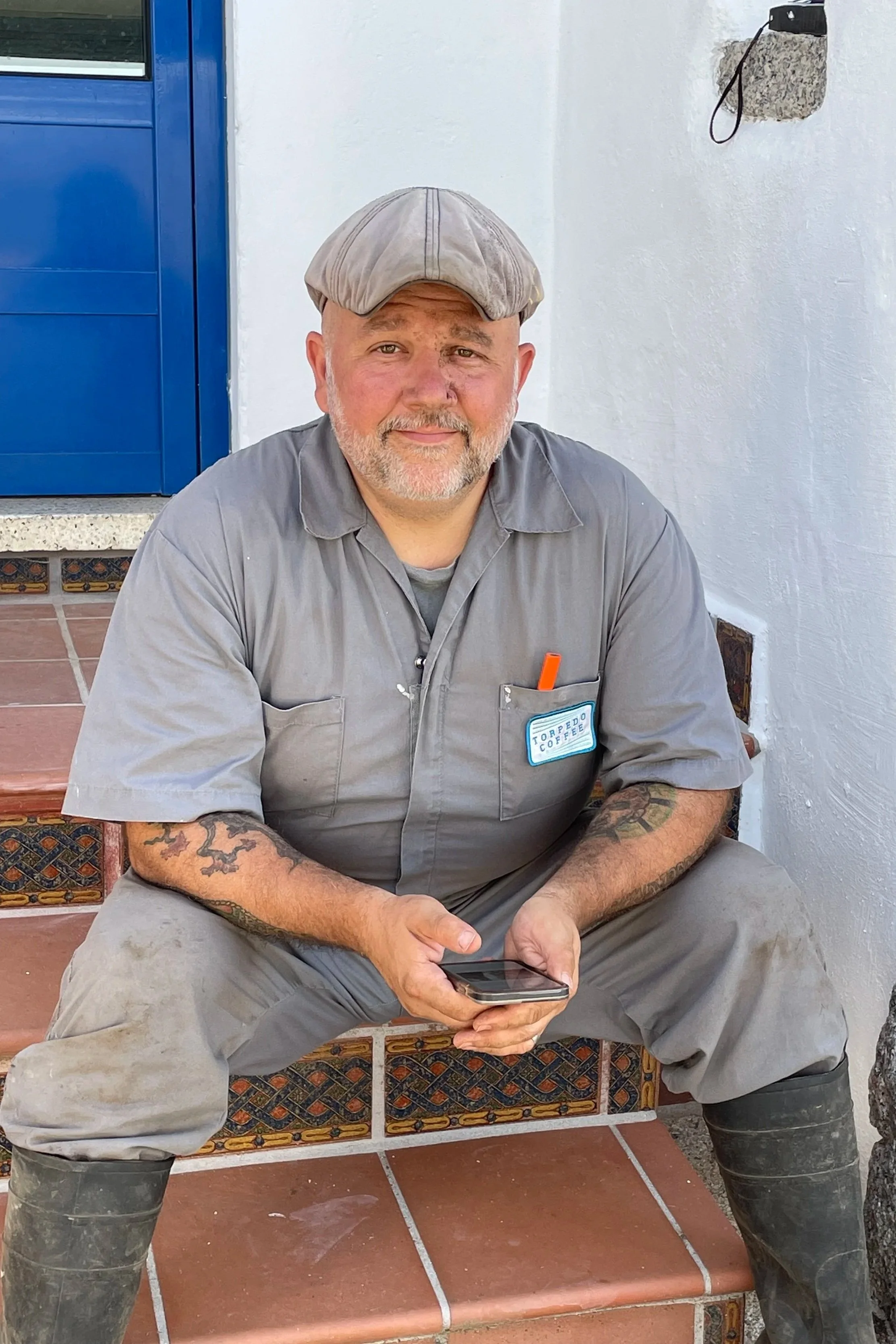 A middle-aged man with tattoos on his arms, wearing a grey uniform with a name tag and a grey cap, sitting on tiled steps outside, holding a smartphone.