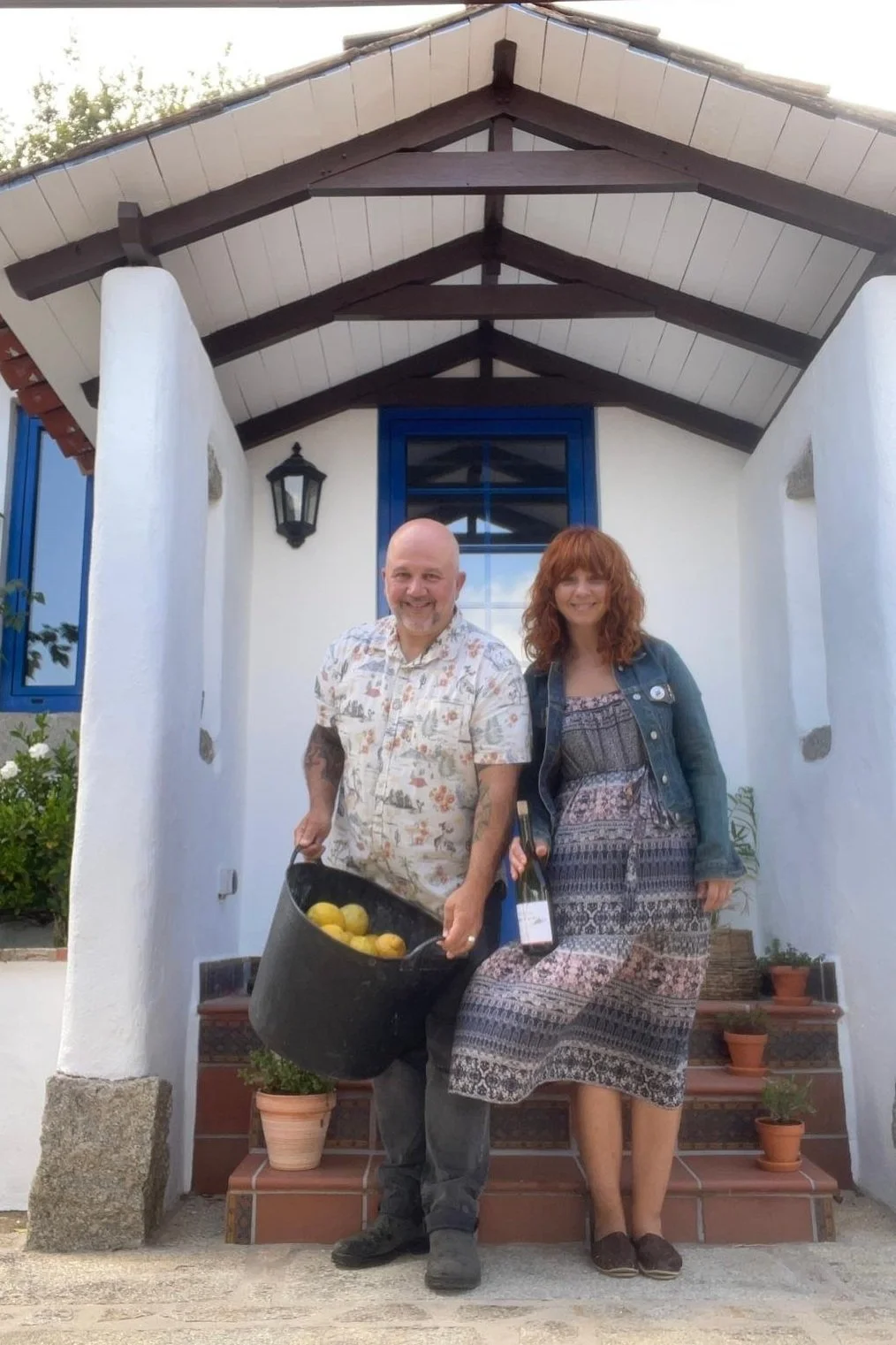 A man and woman standing on front steps of a white house with blue window frames, smiling. The man is holding a bucket of yellow lemons and a bottle of wine. The woman is wearing a patterned dress and denim jacket, with a potted plant on the steps beside her.