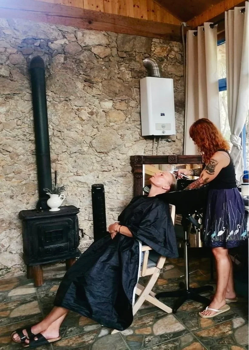 A woman getting her hair washed at a salon, sitting in a chair with a black cape, while hairdresser with red hair and tattoos washes her hair at a sink. The setting has a rustic stone wall, a wood stove, and a window with curtains.