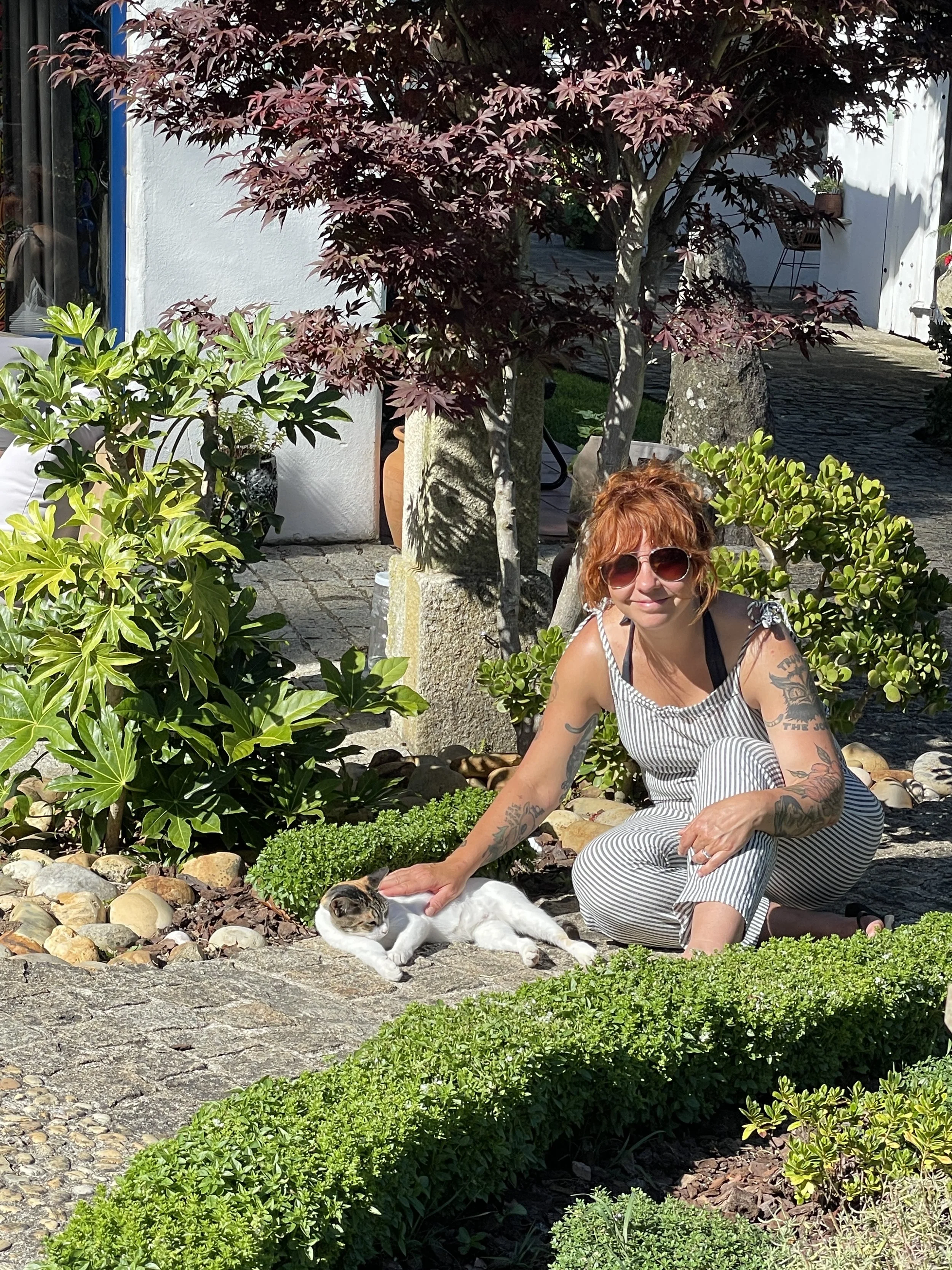 A woman with red hair, sunglasses, and tattoos on her arms, wearing a striped jumpsuit, is sitting on a stone path in a garden. She is petting a white and gray cat lying on the ground. The garden has various green plants, a small shrub, and a tree wi