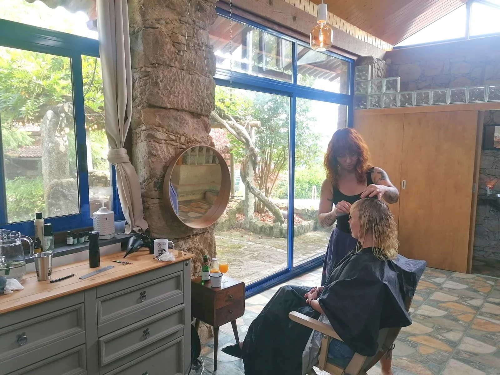 A hairstylist is styling a woman's hair in a modern, rustic salon with large windows, stone walls, and outdoor greenery visible.
