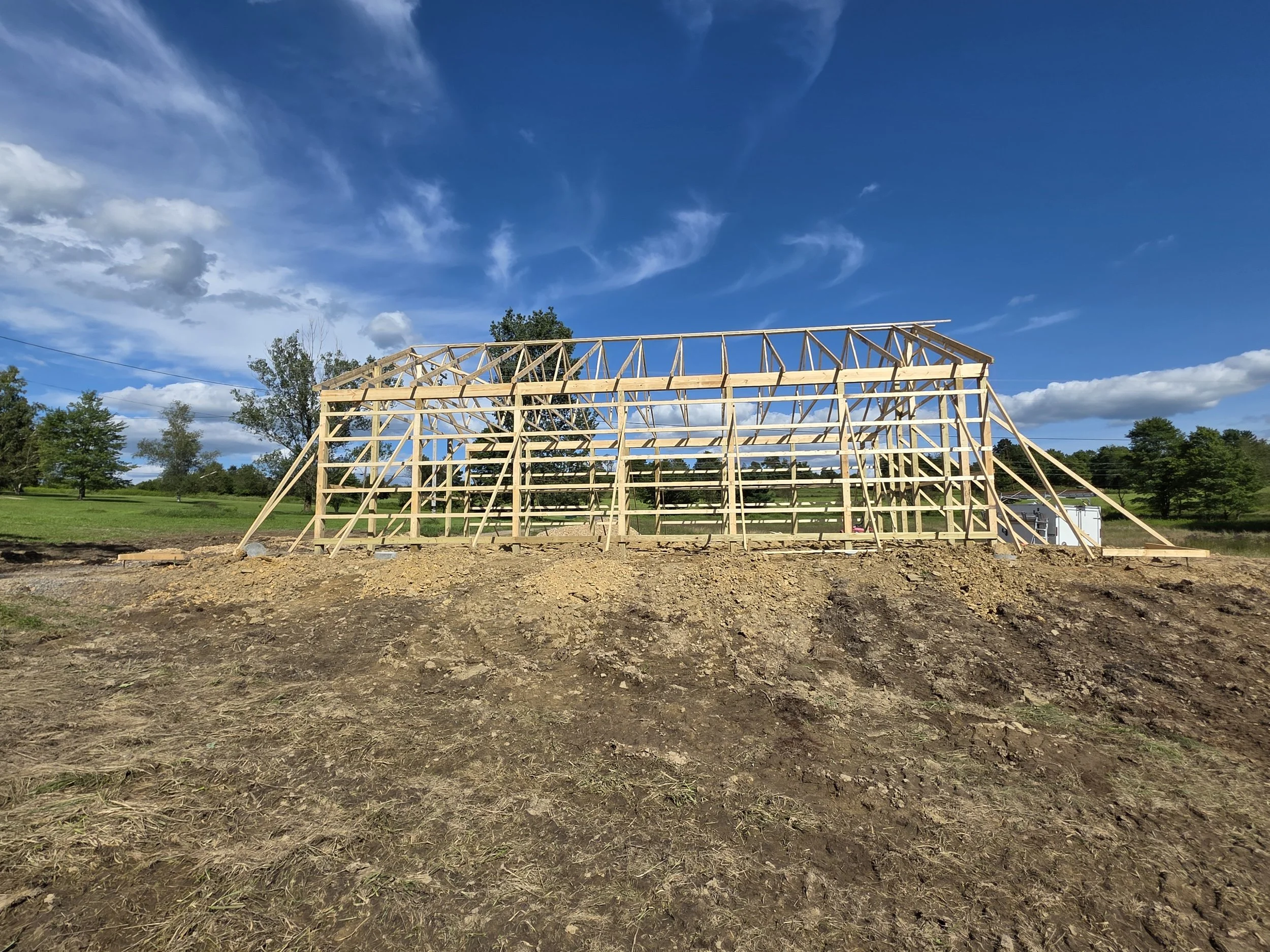 A wooden building frame under construction on a grassy field with trees and a partly cloudy blue sky.