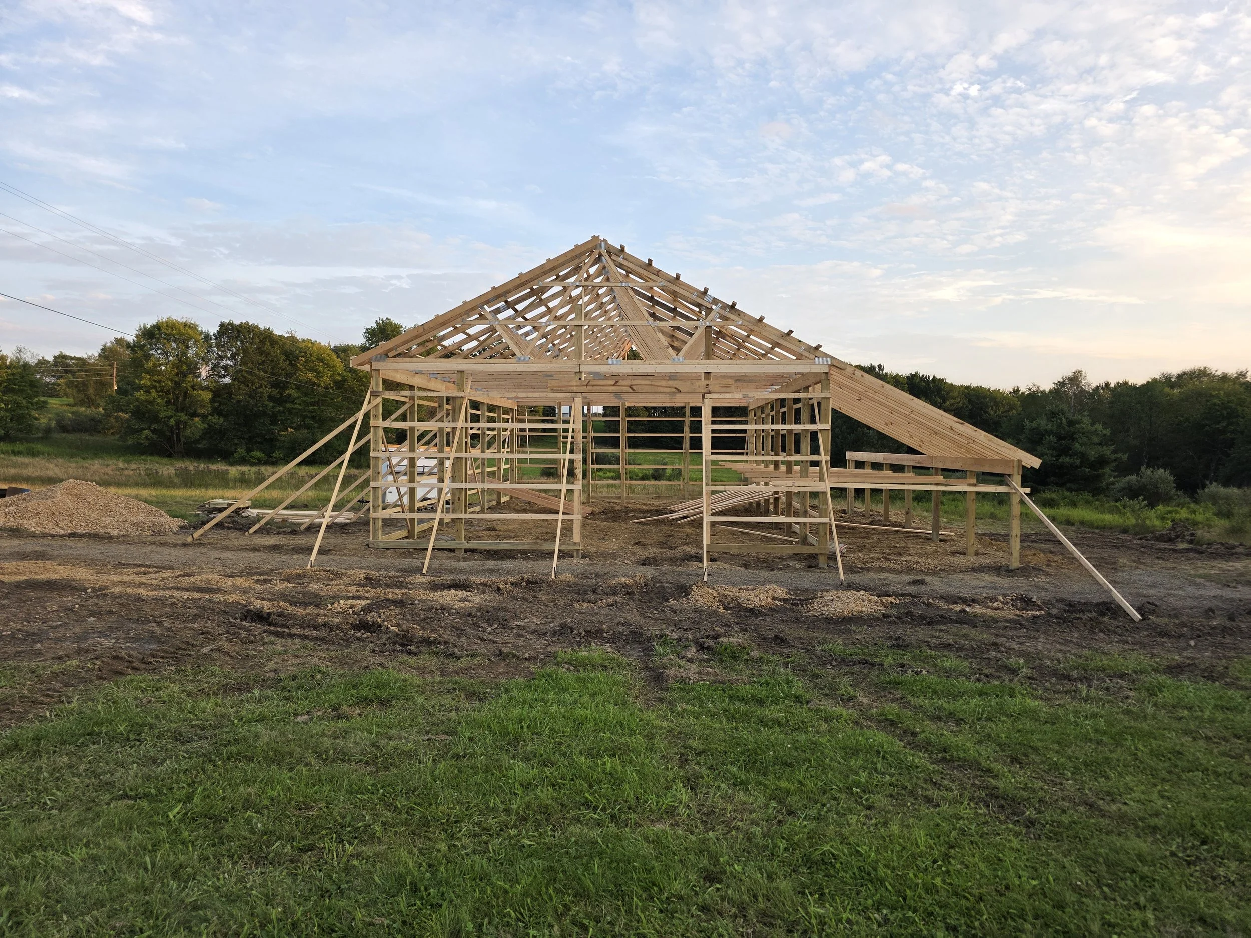 Wooden framework of a house under construction, set against a background of trees and a partly cloudy sky at sunset.