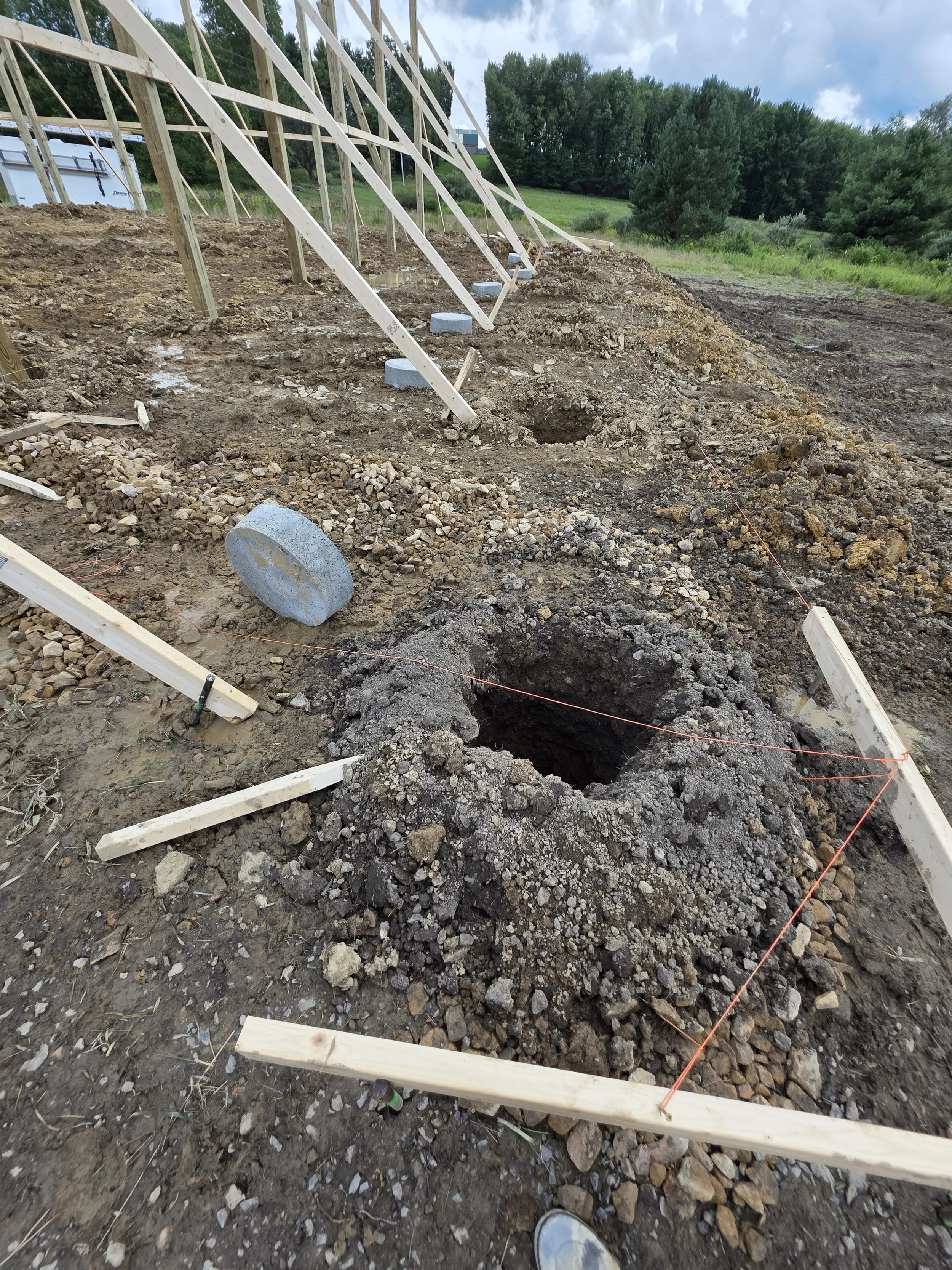 Construction site with wooden framework and drilled holes in the ground, surrounded by dirt, rocks, and construction markings, with trees and cloudy sky in the background.