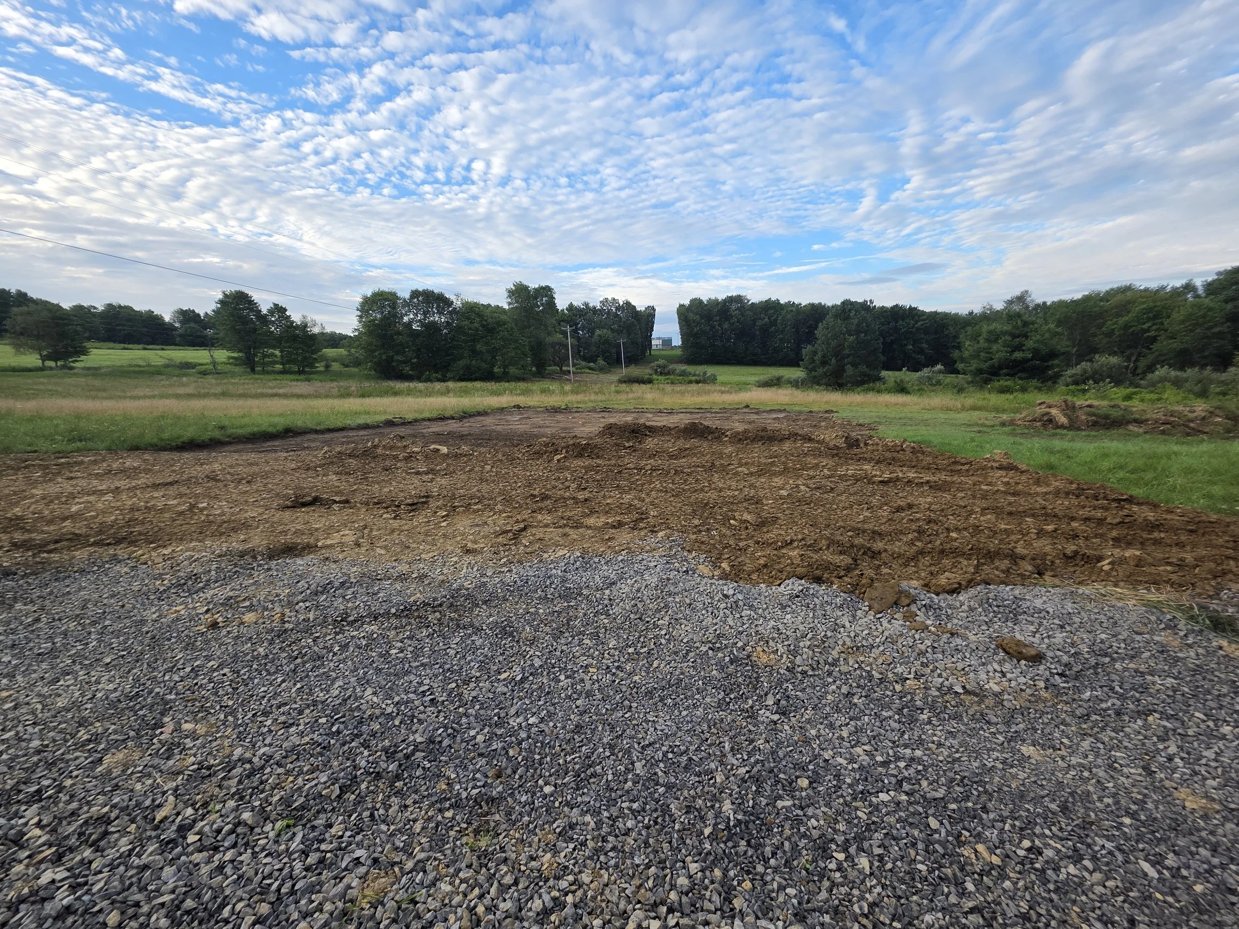 View of a rural landscape with a partly cloudy sky, green trees, and a dirt and gravel foreground.