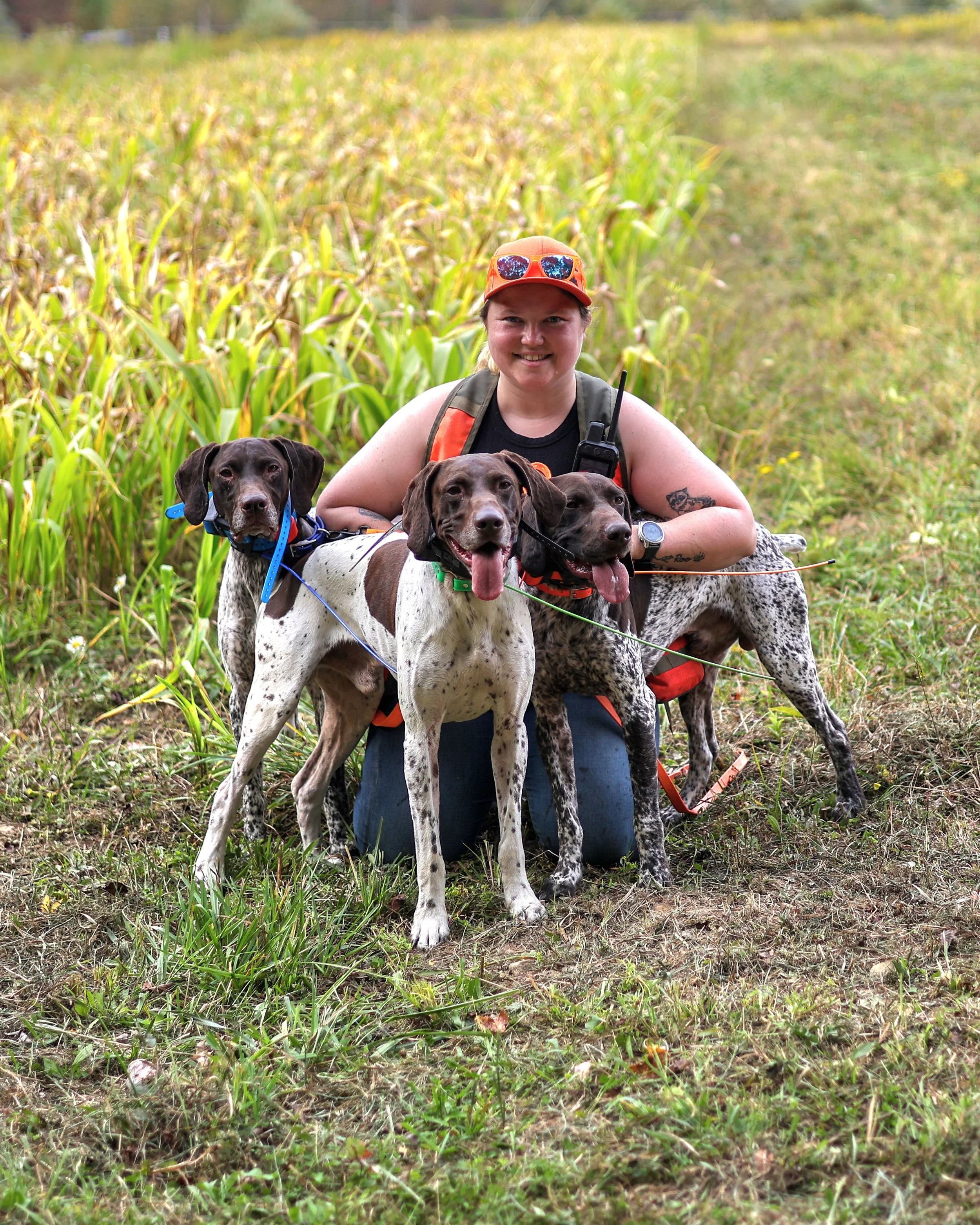 A woman crouching in a grassy field with four German Shorthaired Pointers puppies. She is wearing an orange cap with sunglasses on top, a black shirt, and has a tattoo on her left arm. The puppies are standing in front of her, and the background shows a field of tall green plants with a blurry tree line.