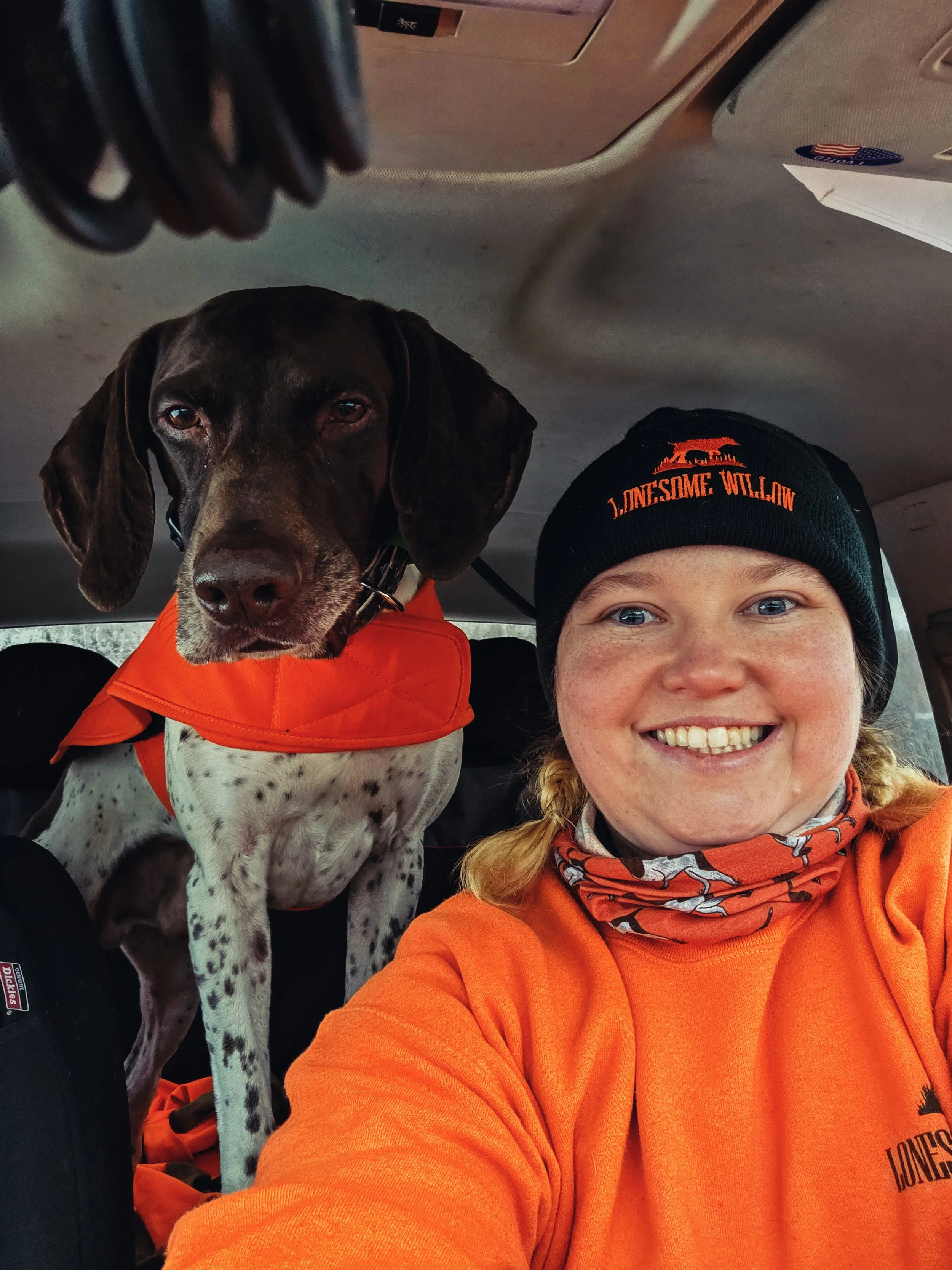 A woman wearing an orange shirt, a black beanie that says "Lonesome Willoughby," and a bandana around her neck, smiling inside a vehicle. Next to her is a German Shorthaired Pointer dog with brown and white fur, wearing an orange vest or jacket. The dog is sitting on the vehicle seat and is looking into the camera along with the woman.