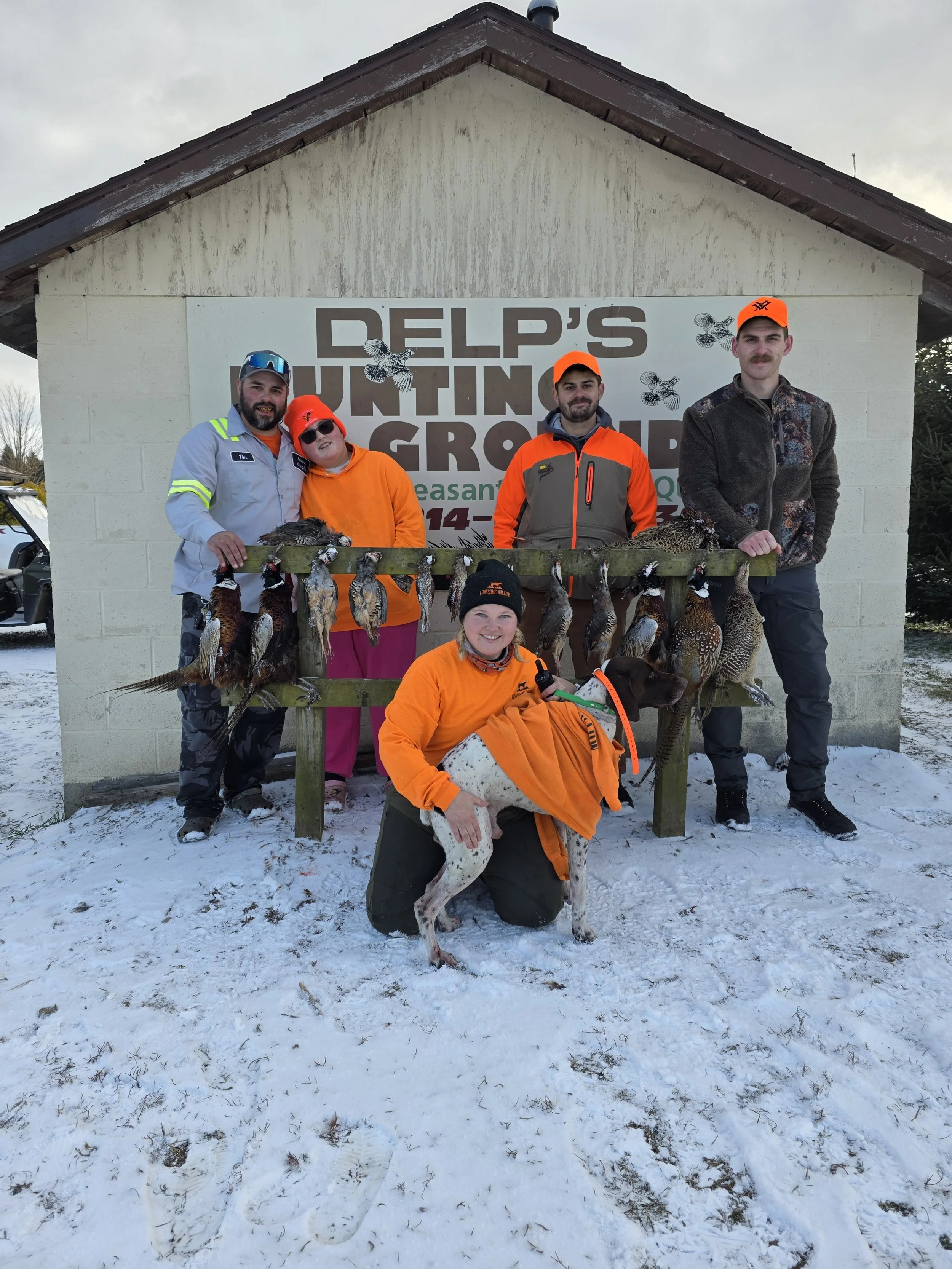 Four adults and a young girl posing with a large hunting dog and a rack of hunted birds in front of a building with a sign that reads 'Delp's Hunting Ground'. The group is dressed in outdoor hunting and hunting gear, and the scene is snowy and outdoors.