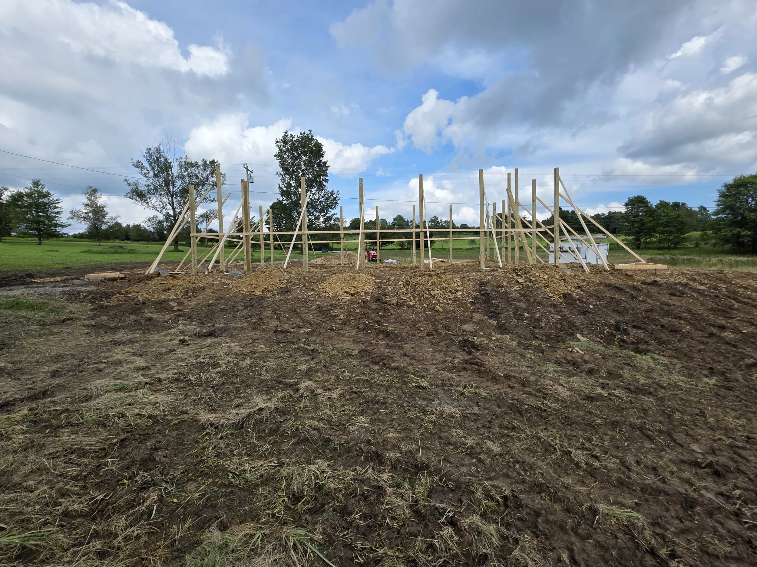 Construction site with wooden framework for a building, surrounded by grass and trees, under a partly cloudy sky.