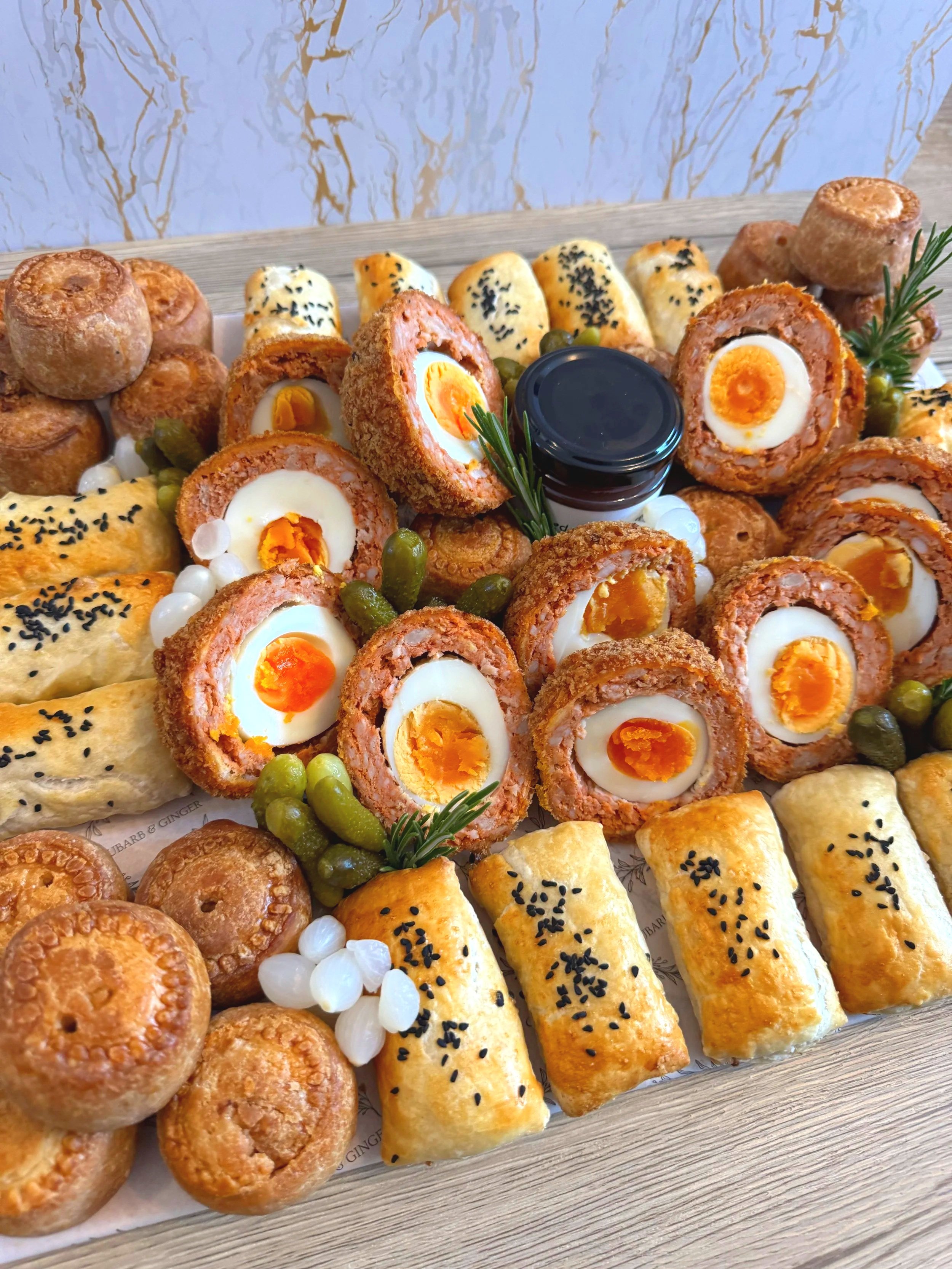 A platter of assorted savory pastries, including Scotch eggs, sausage rolls, and sausage muffins, garnished with green grapes, onions, and sprigs of rosemary, with a small jar of sauce in the center.