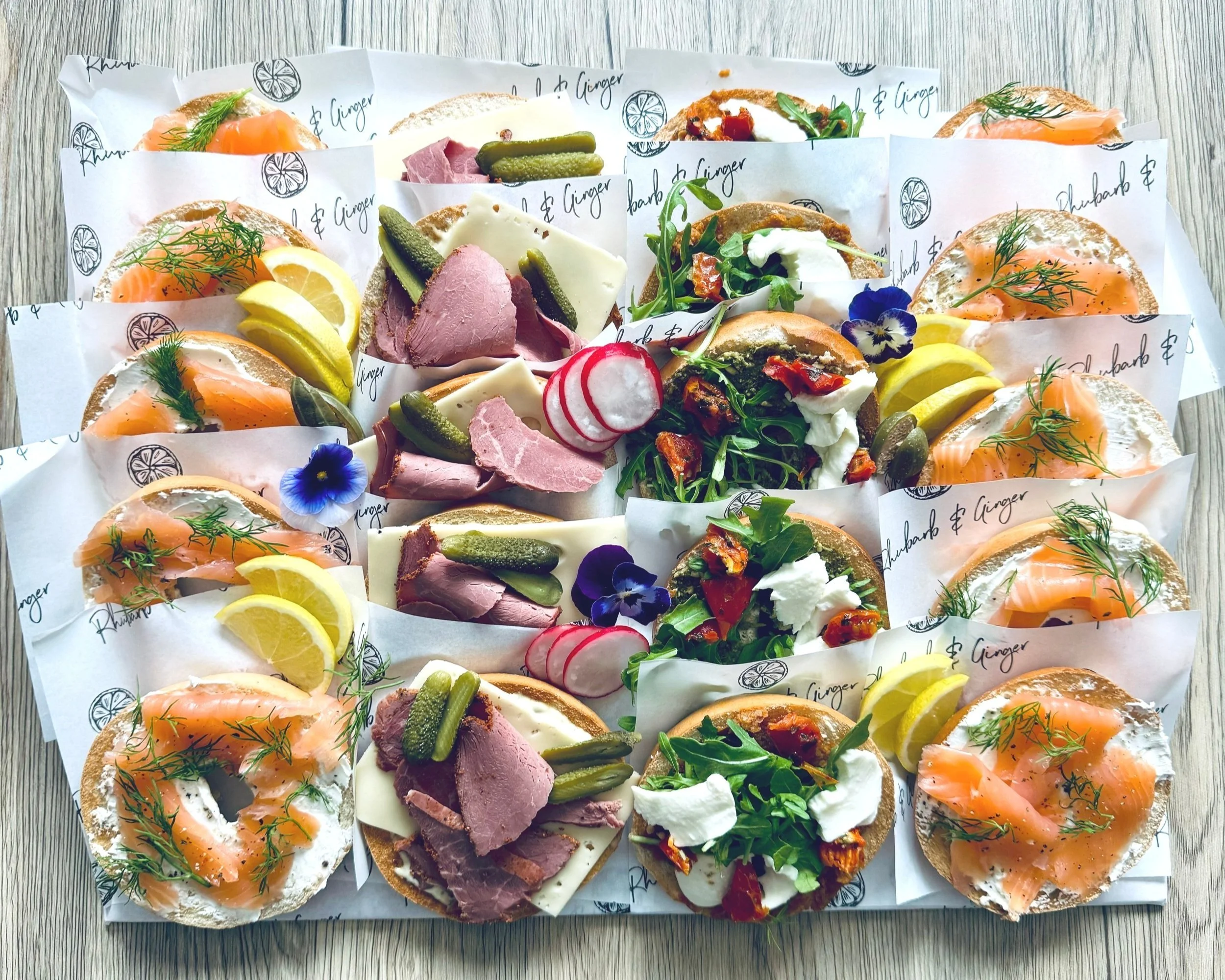 A platter of assorted open-faced sandwiches with smoked salmon, cured meats, cheeses, and various garnishes, arranged on individual pieces of branded parchment paper on a wooden surface.
